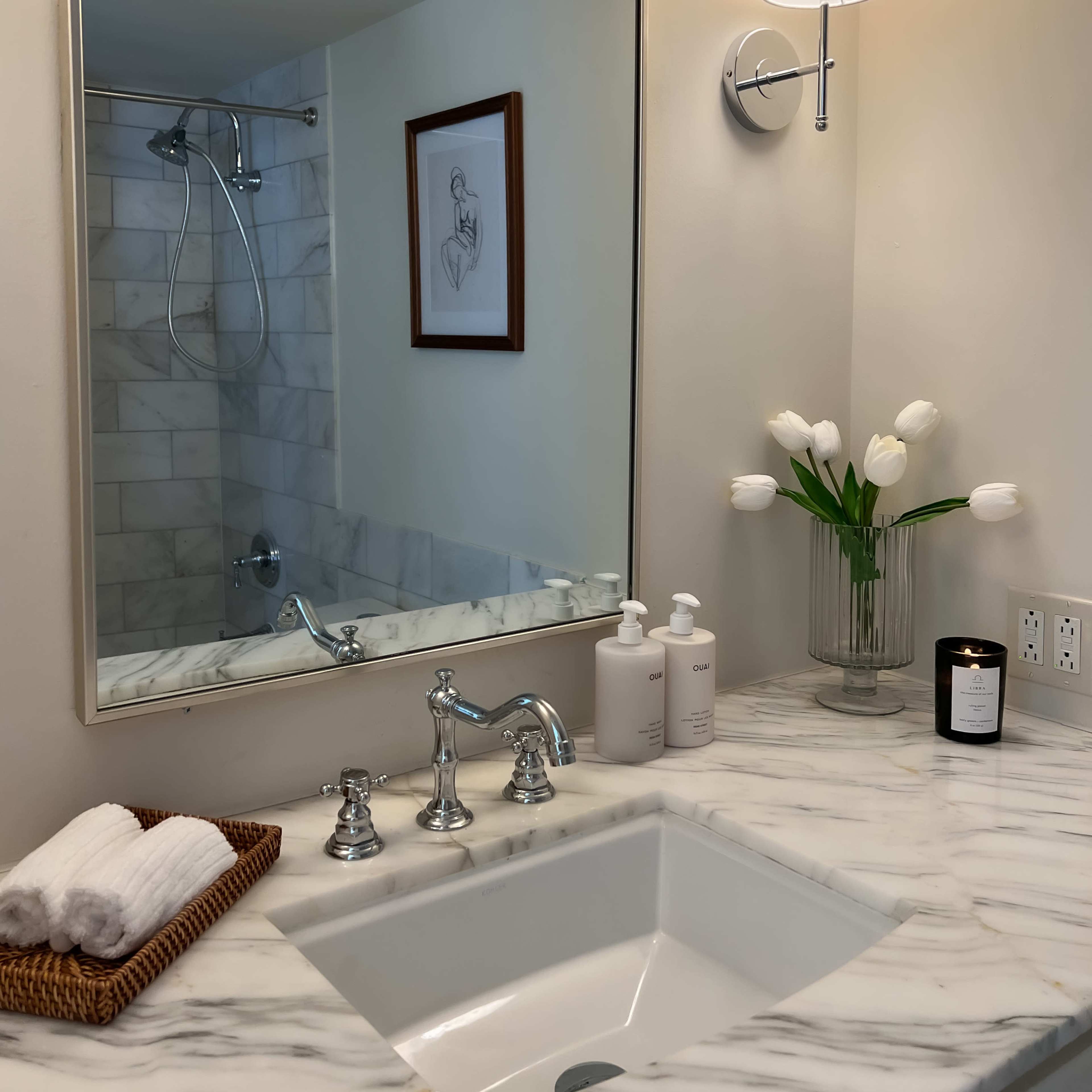 A modern bathroom featuring a marble countertop with a sink, a wall-mounted light fixture, a mirror, and a vase of tulips.