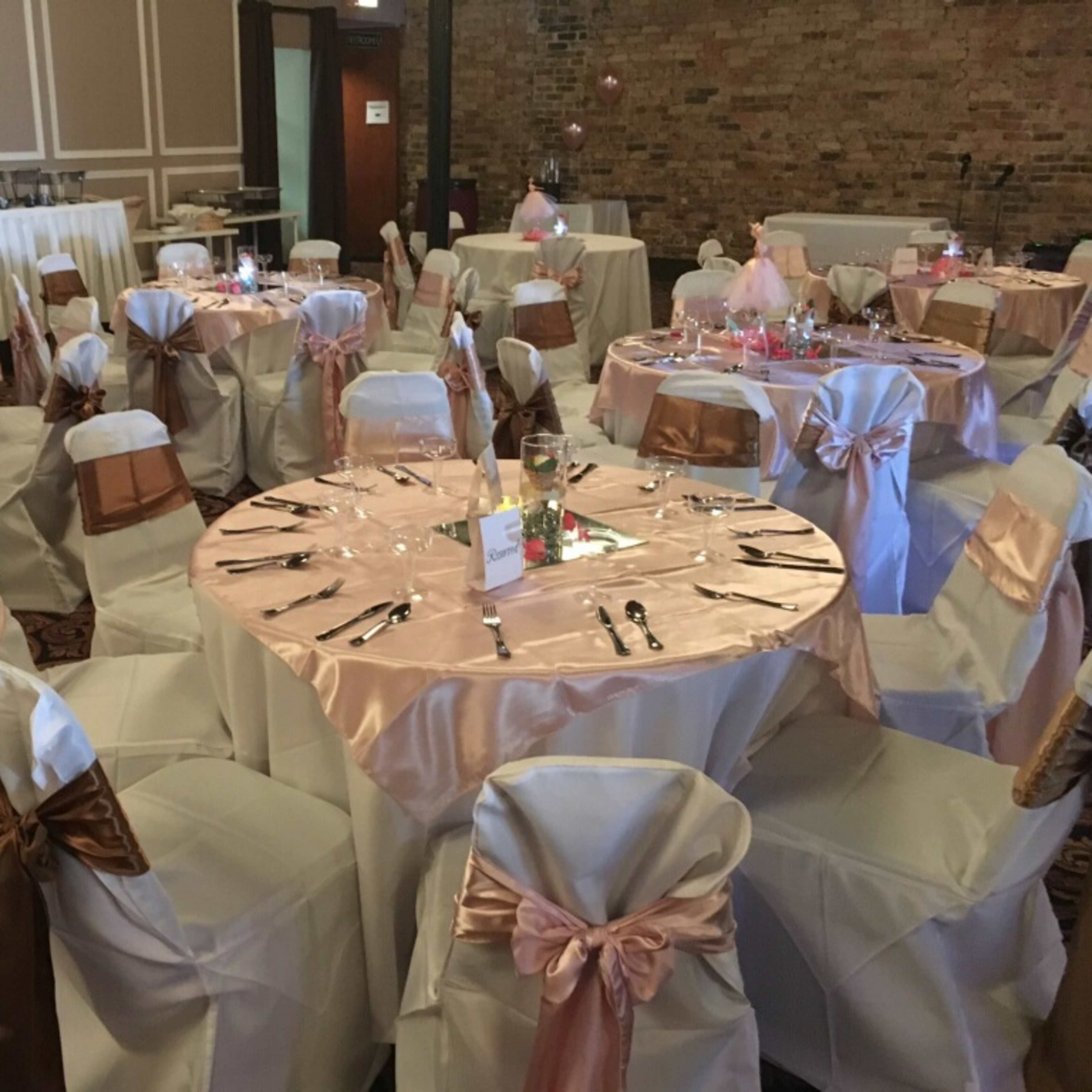 A banquet hall set up with multiple round tables covered in light pink tablecloths and decorated with bows in a brown hue, all arranged with place settings and centerpieces.