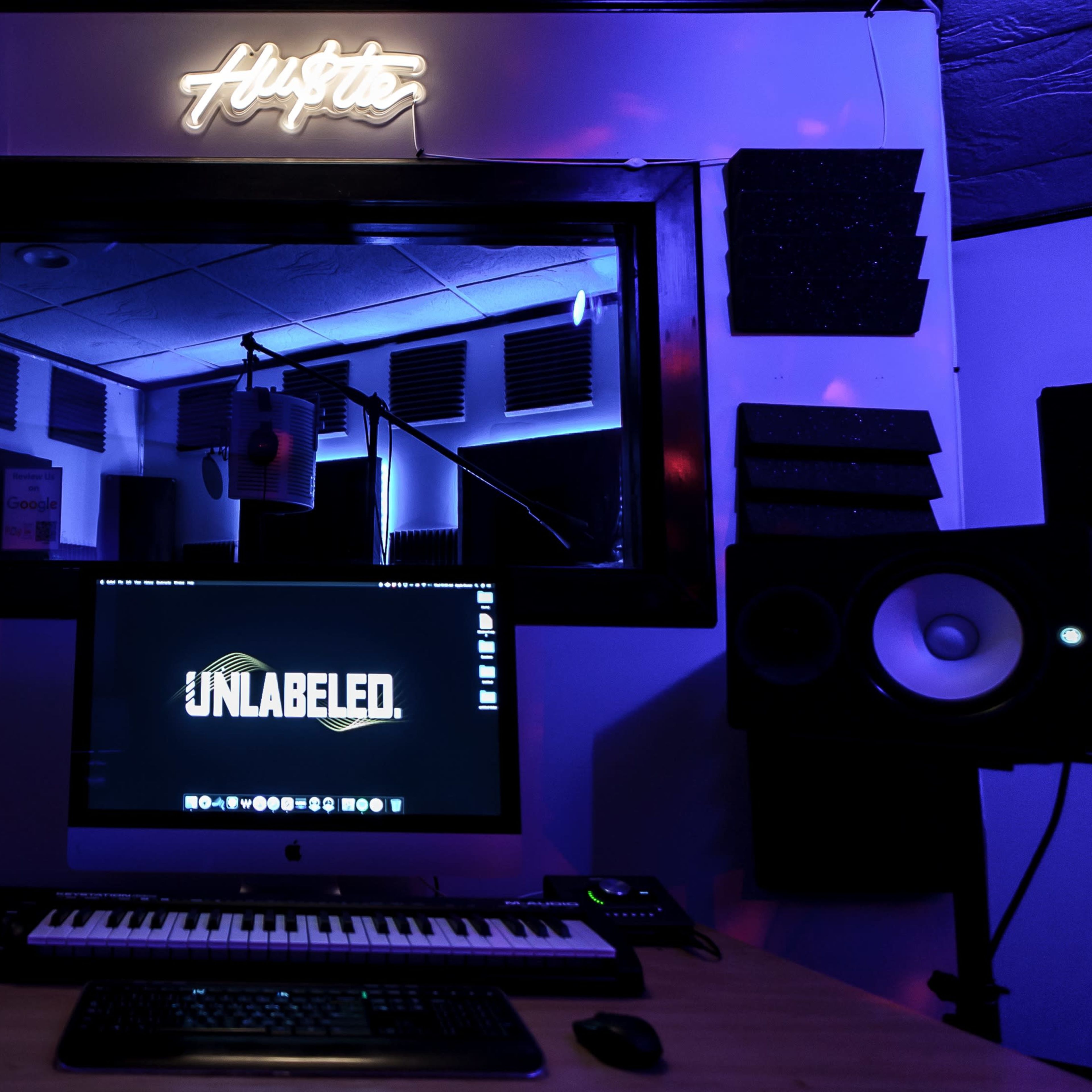 A music production studio featuring a computer, keyboard, and speakers, with a microphone setup visible in the background and neon lighting.