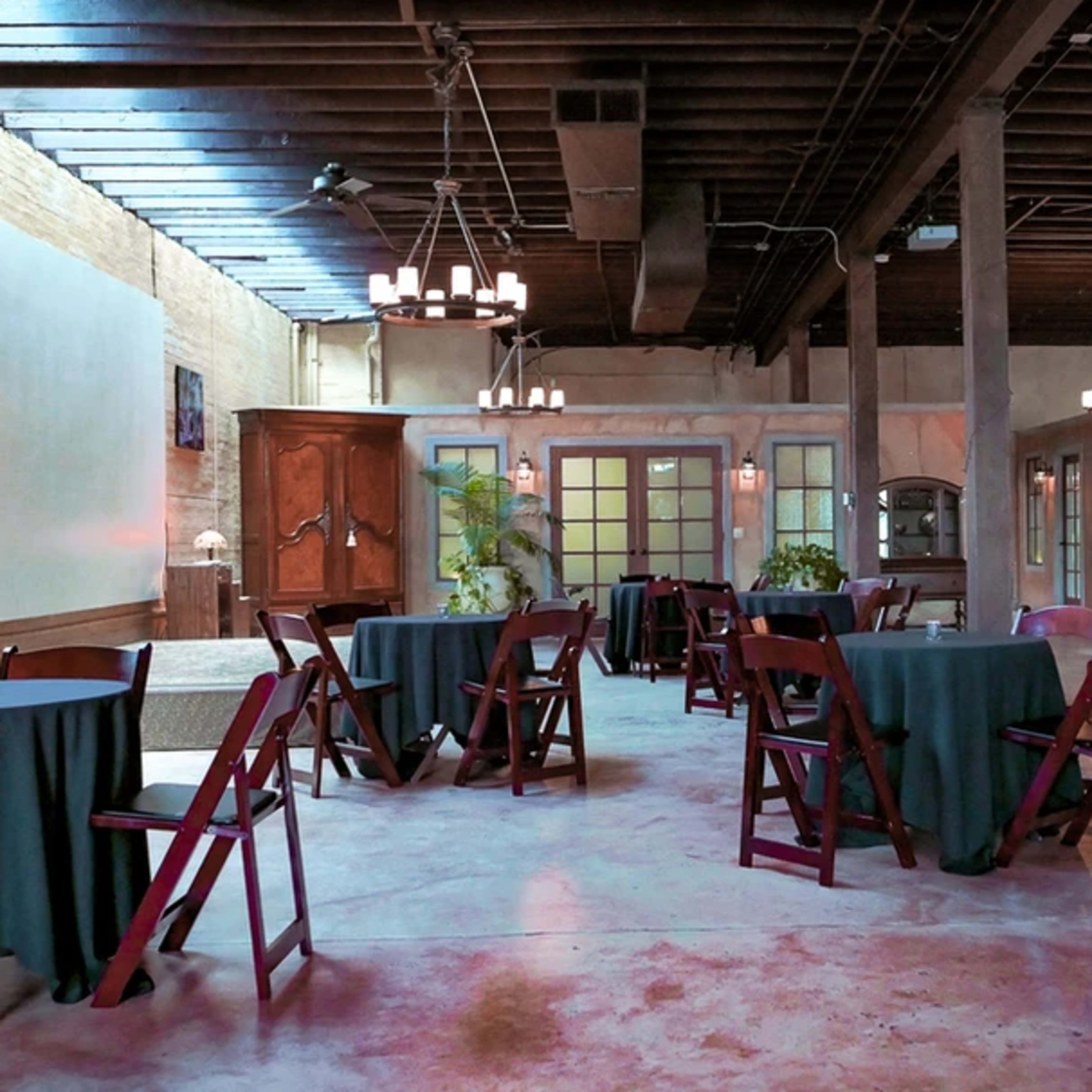 The interior of a spacious venue features several round tables with dark tablecloths surrounded by wooden chairs, illuminated by dim lighting and adorned with chandeliers.