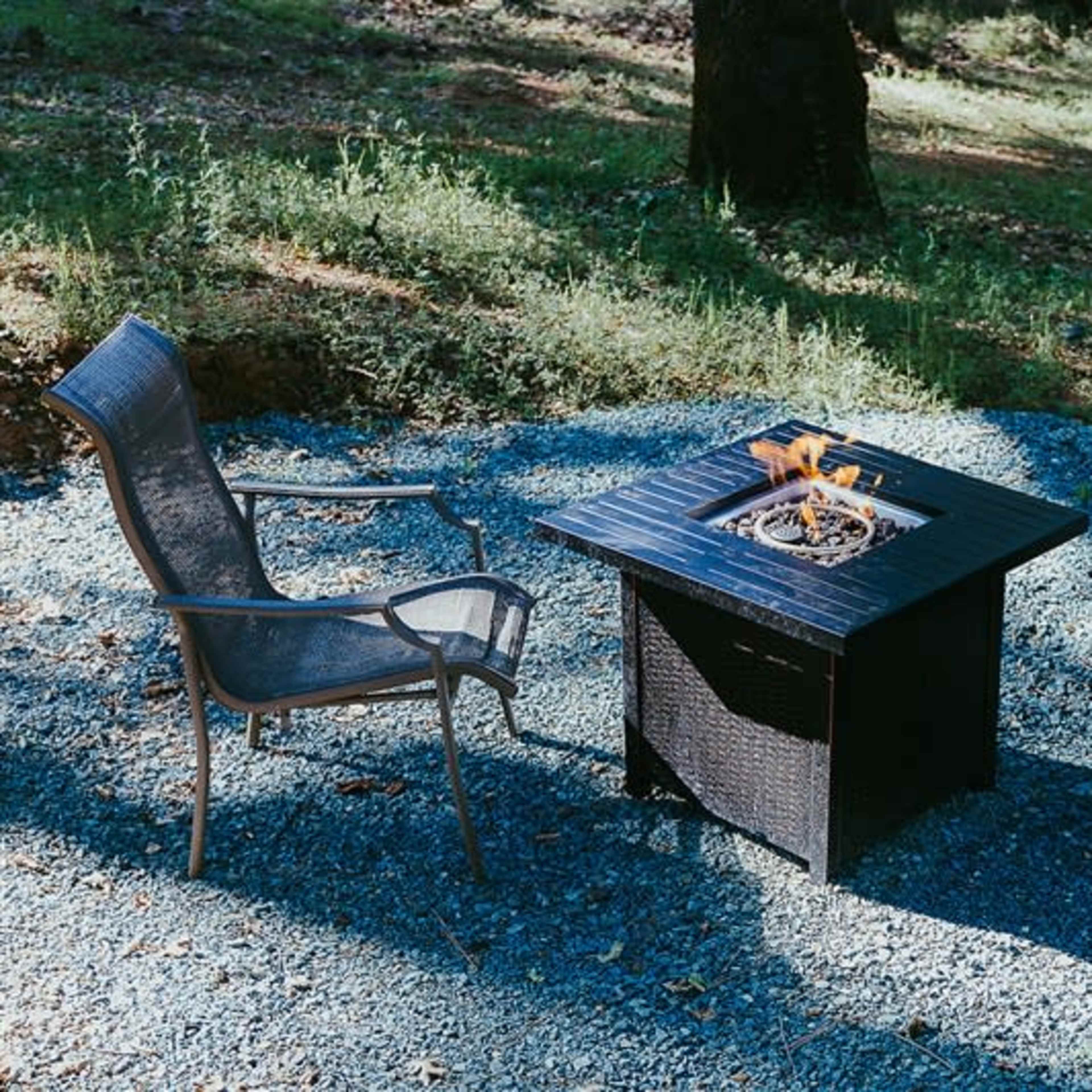 A fire pit surrounded by two chairs is set on a gravel surface in a wooded area.
