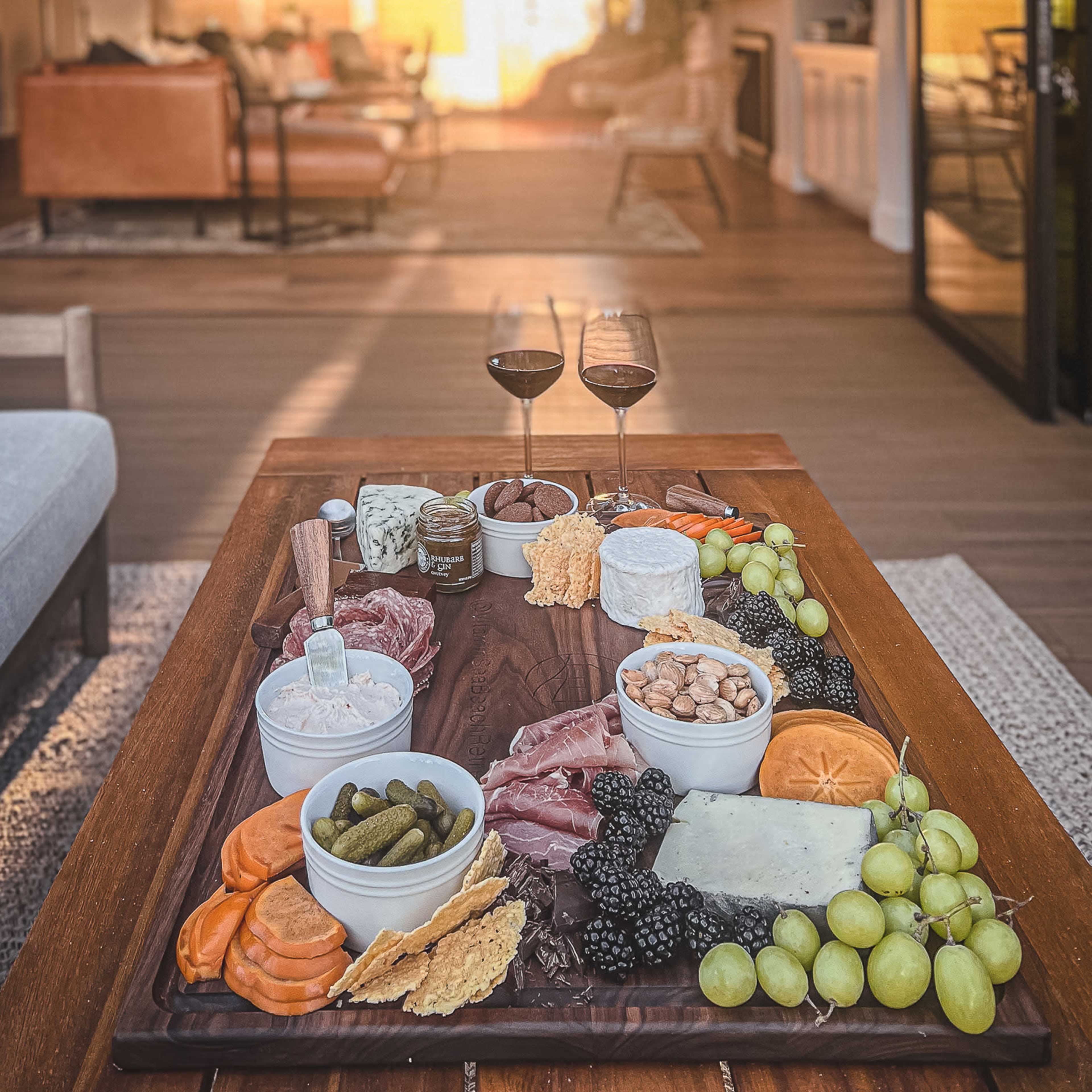 A wooden table is adorned with a charcuterie board featuring assorted meats, cheeses, fruits, and two glasses of red wine, with a cozy living room visible in the background.