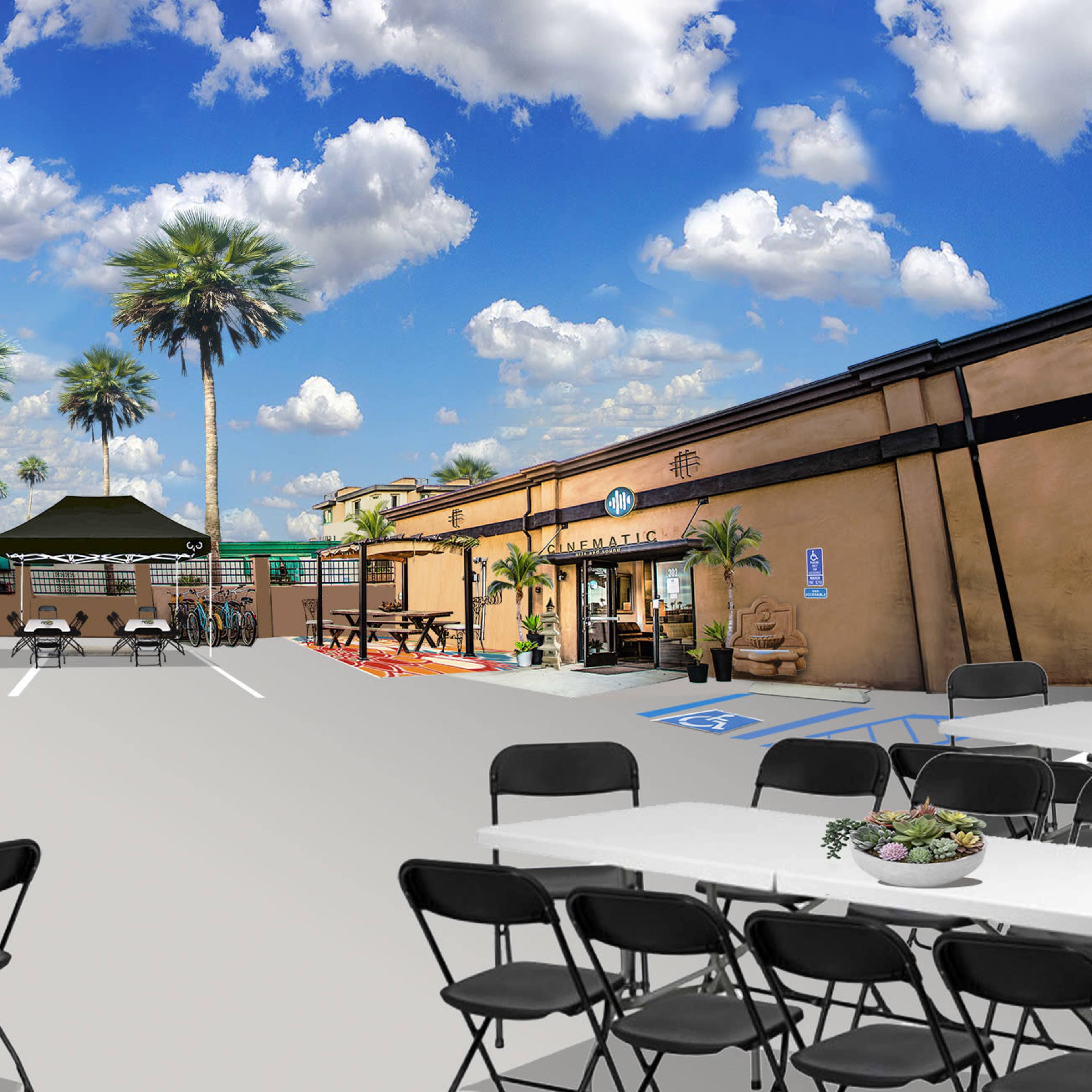 An outdoor event space with tables and black chairs arranged under tents, surrounded by palm trees against a blue sky with clouds.