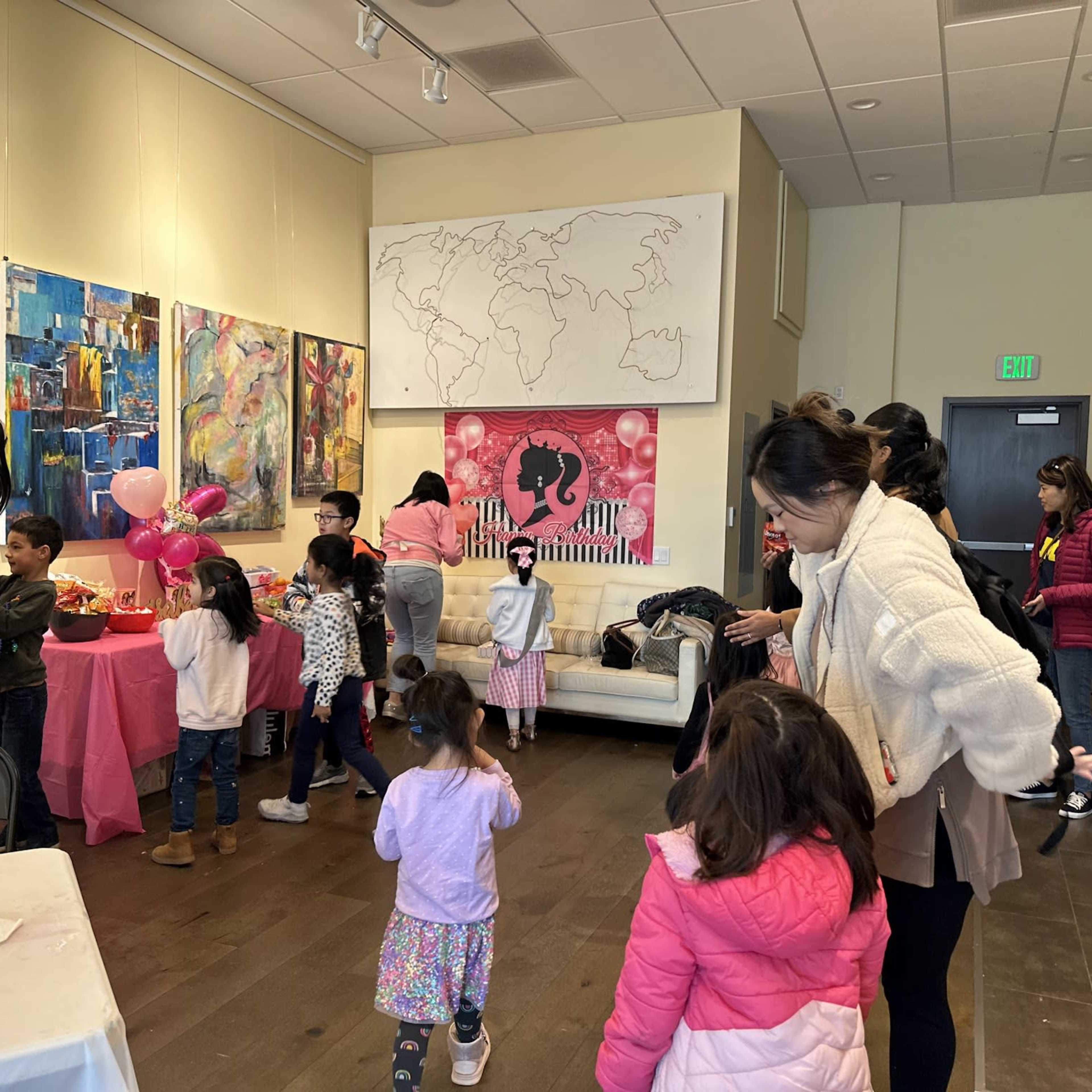 A group of children and adults gather inside a brightly lit room decorated for a party, with pink-themed decorations and artwork on the walls.