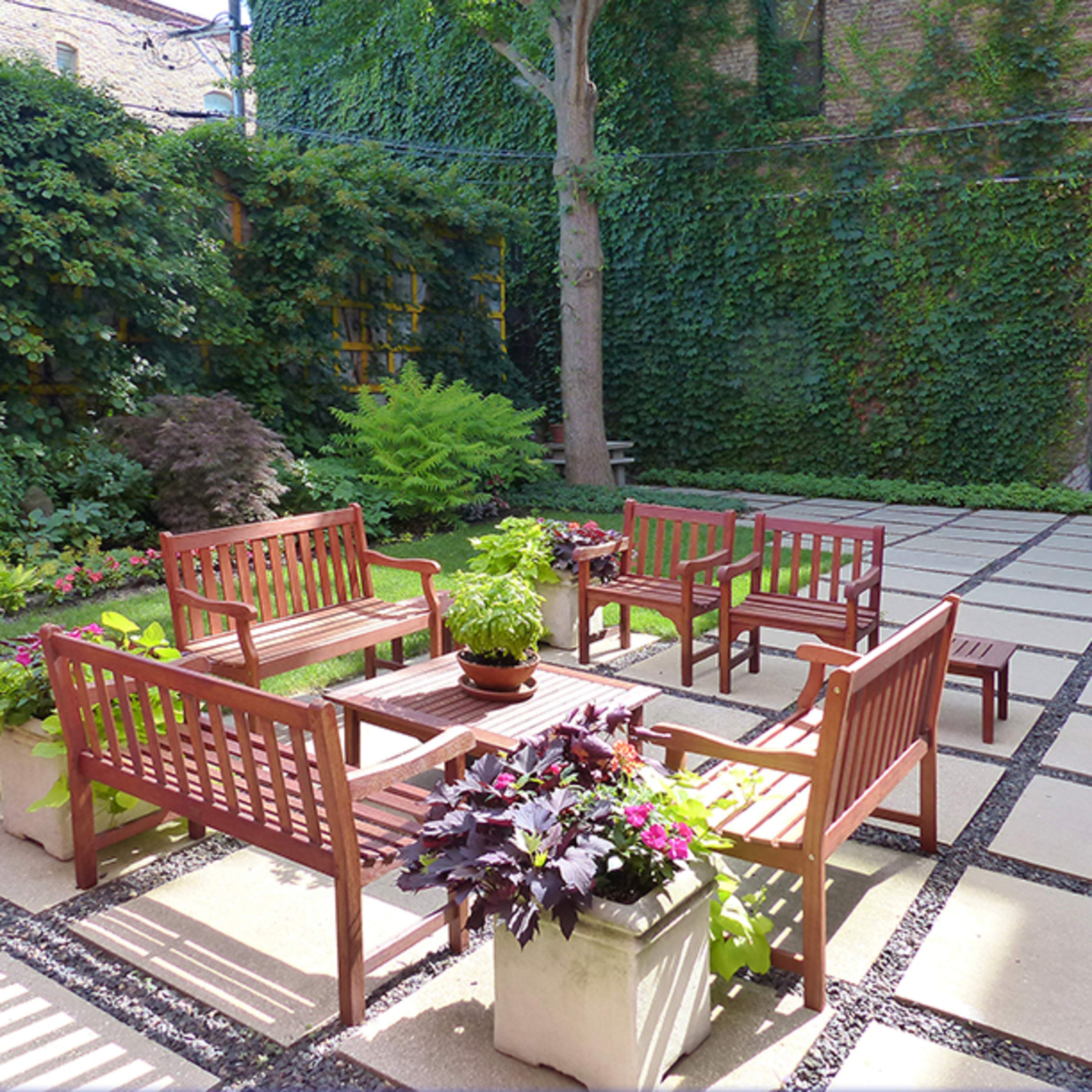 A sheltered courtyard features wooden benches arranged around a central table, surrounded by lush greenery and potted plants.