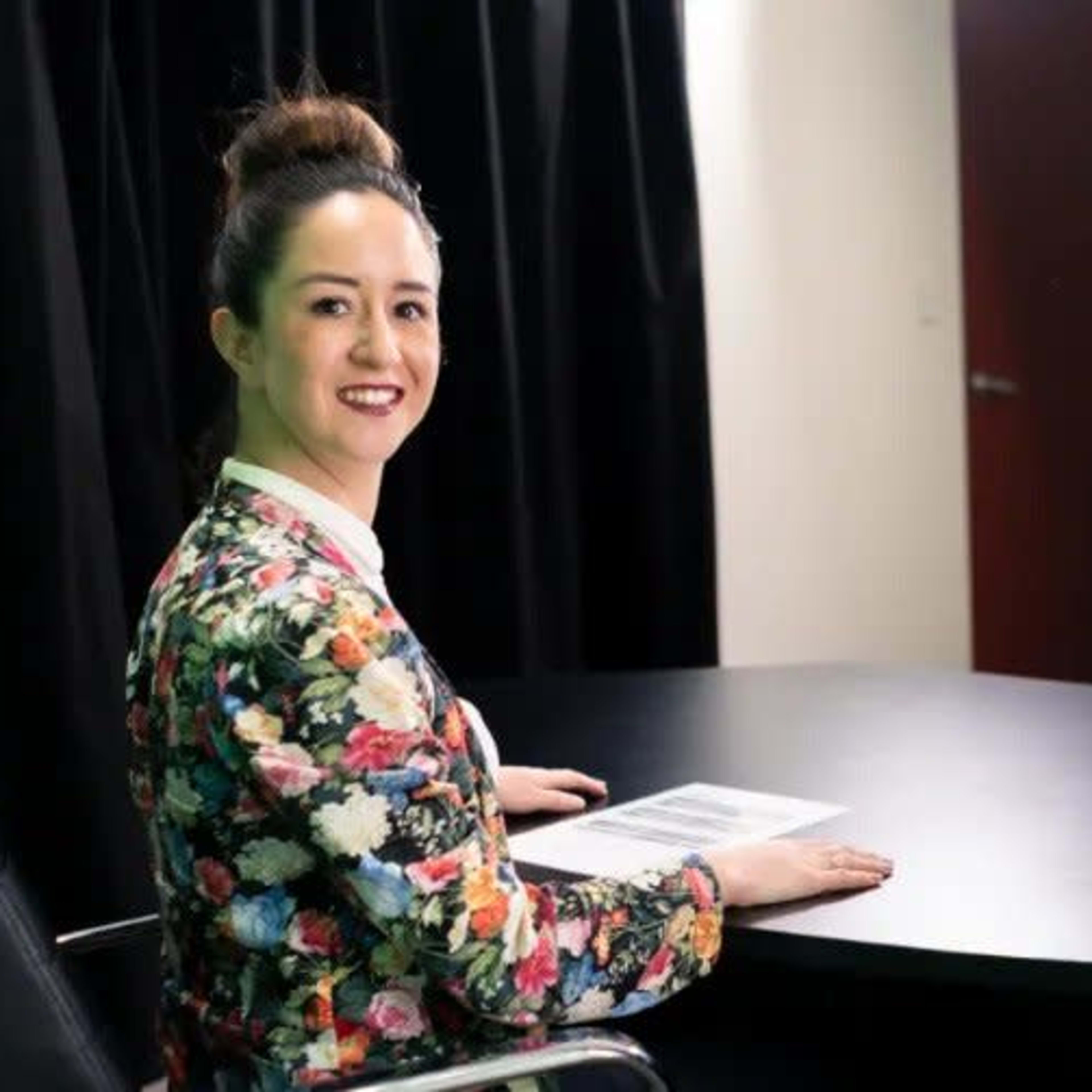 A woman sits at a black table in a studio setting, smiling while looking at the camera, with a presentation displayed on a screen behind her.