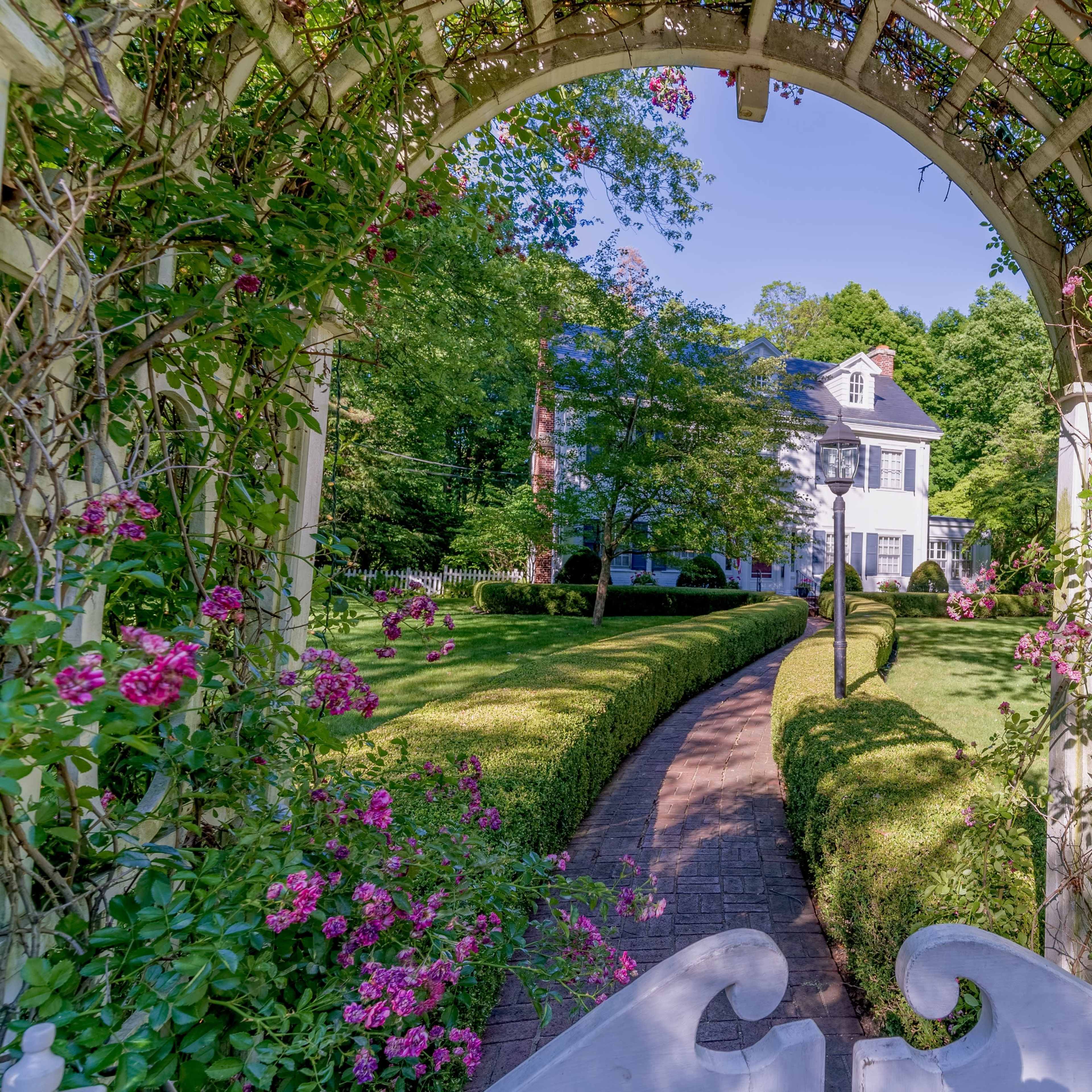 A white house with a black roof is framed by a wooden arbor covered in pink flowers, leading to a brick pathway through a manicured lawn.