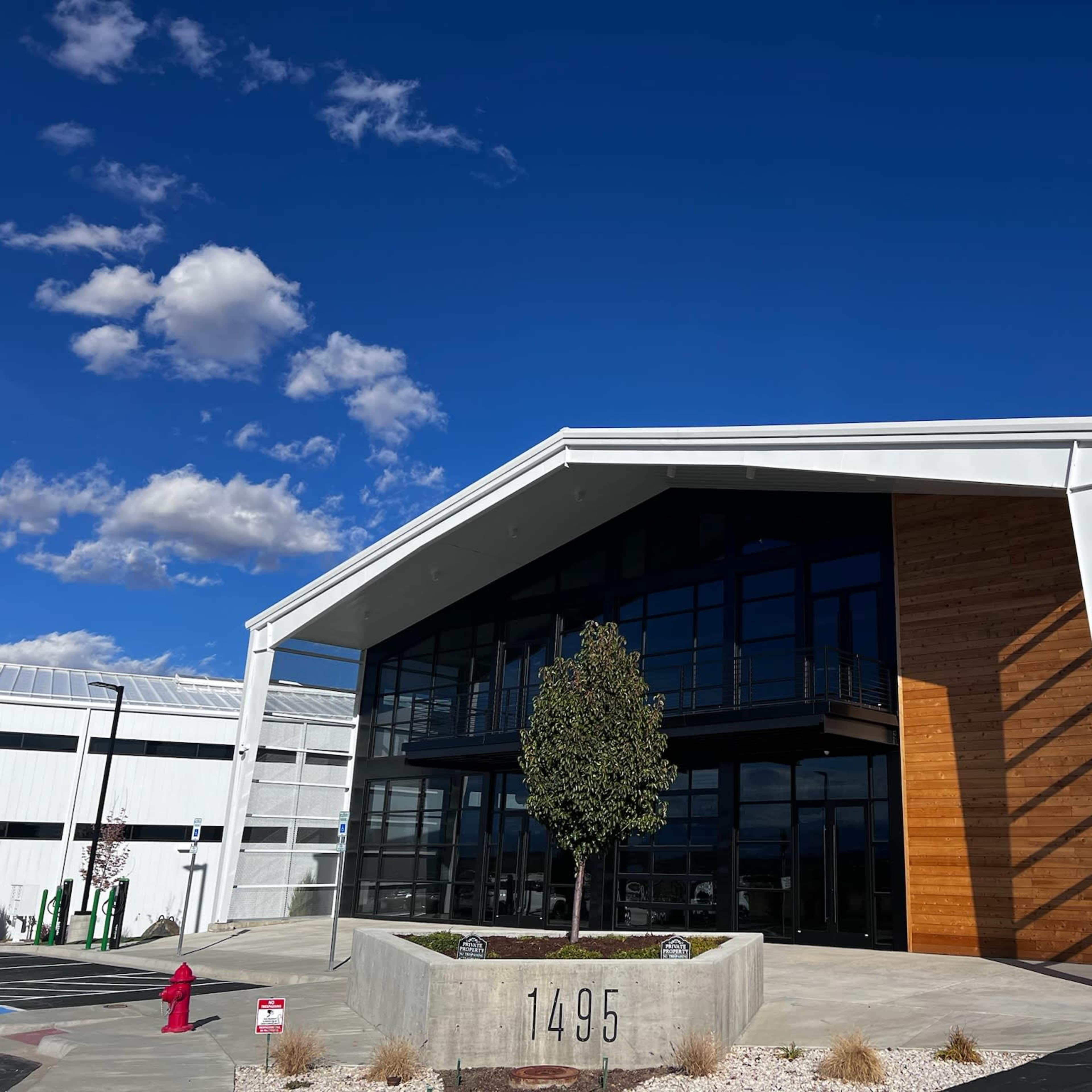 A modern building with a large glass entrance and wooden accents, numbered 1495, is set against a blue sky with scattered clouds.