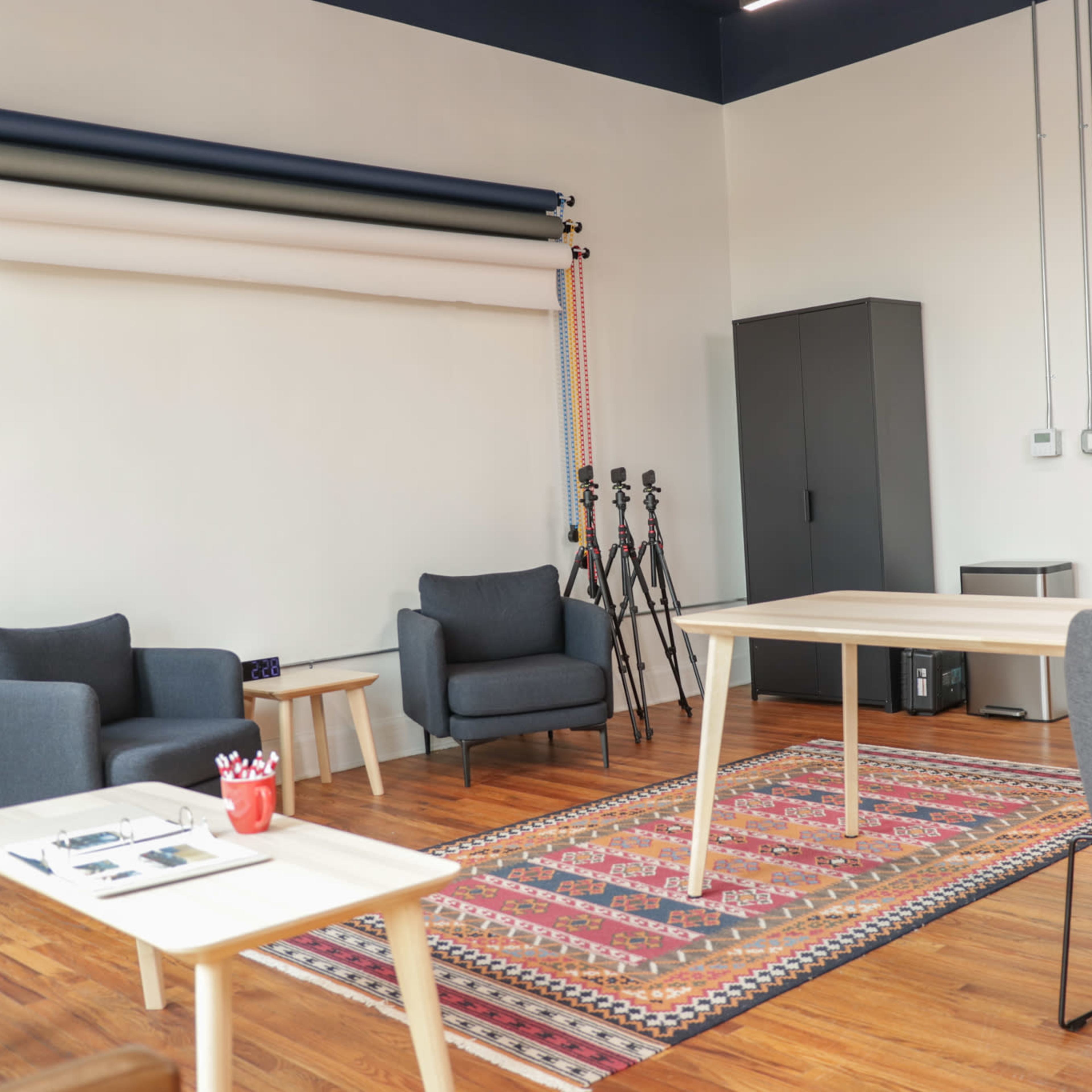 A modern studio with two chairs, a table, a patterned rug, and audio recording equipment, against a backdrop of colored backdrop rolls and a storage cabinet.