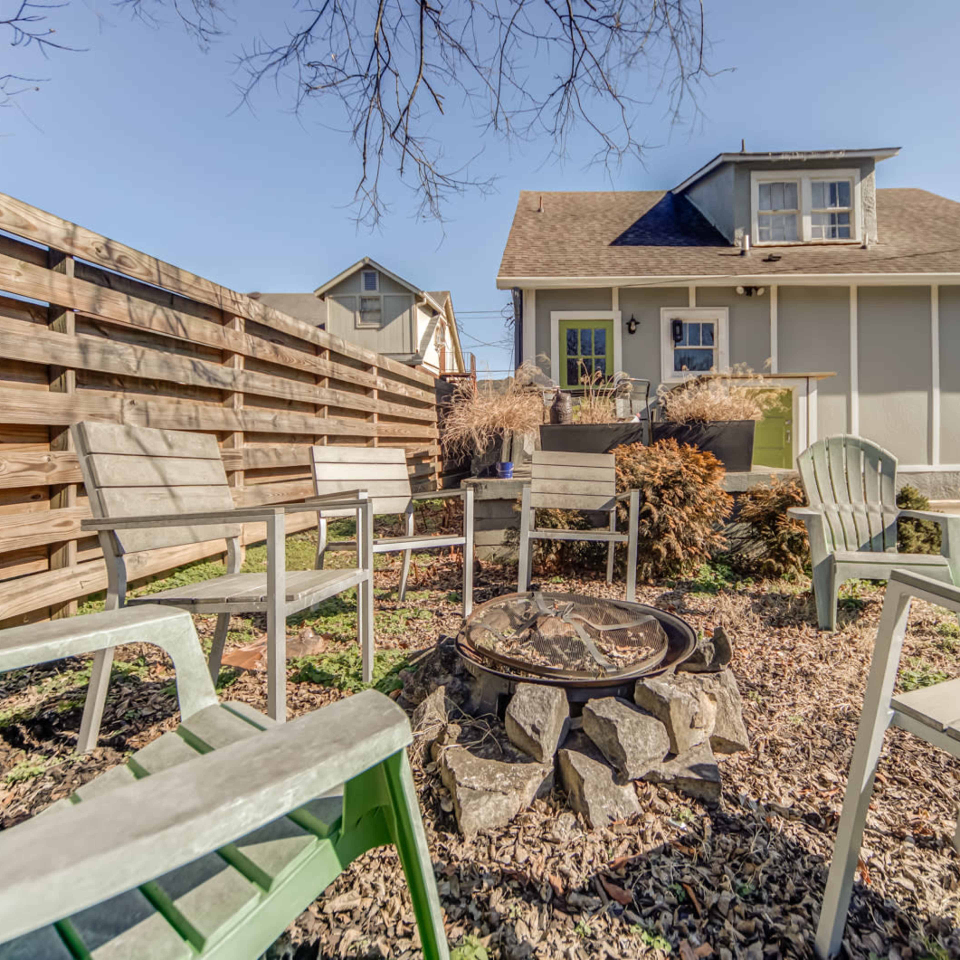 A fire pit surrounded by several chairs is located in a backyard next to a house with a wooden fence.