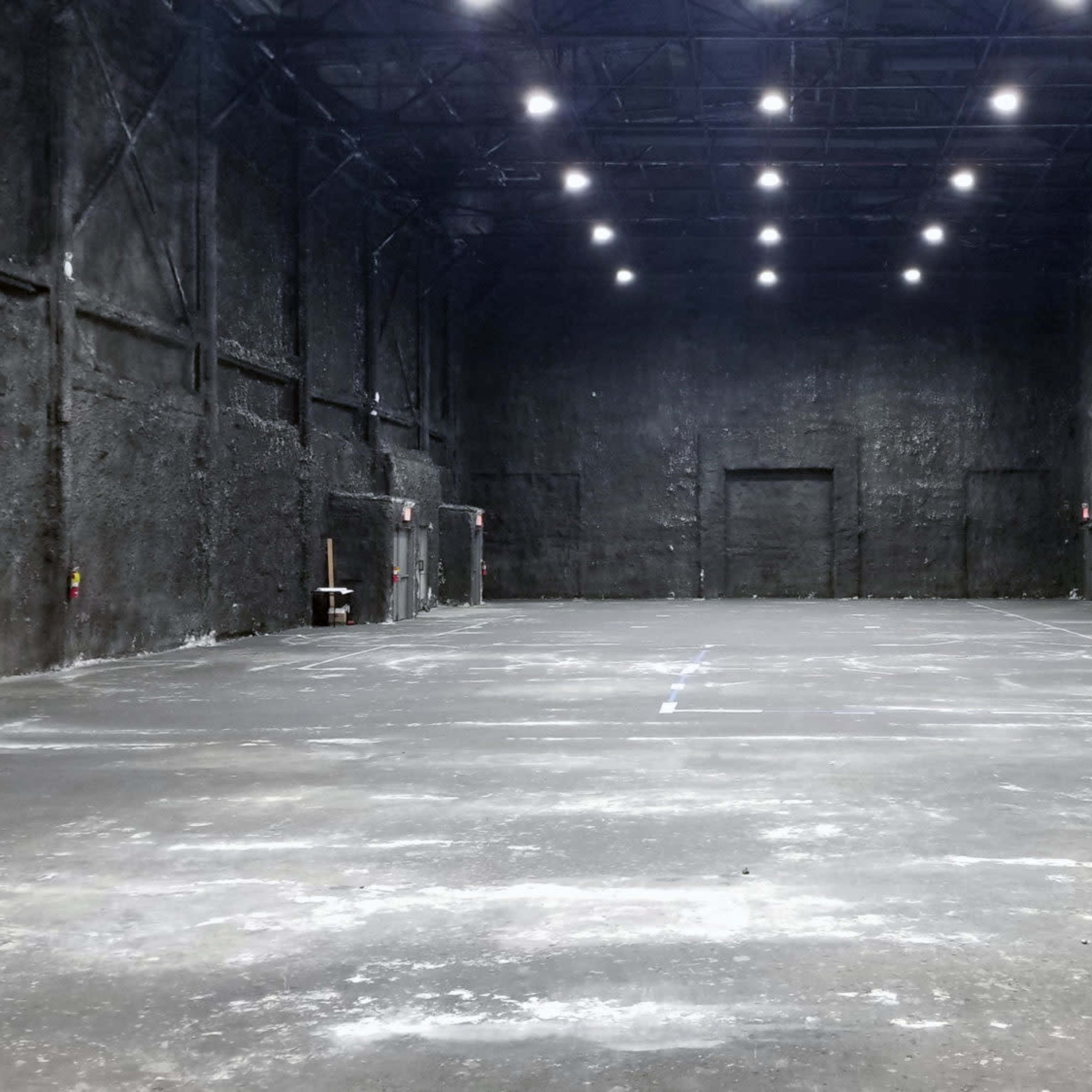 A large, empty industrial space with dark, textured walls and a concrete floor under bright overhead lighting.