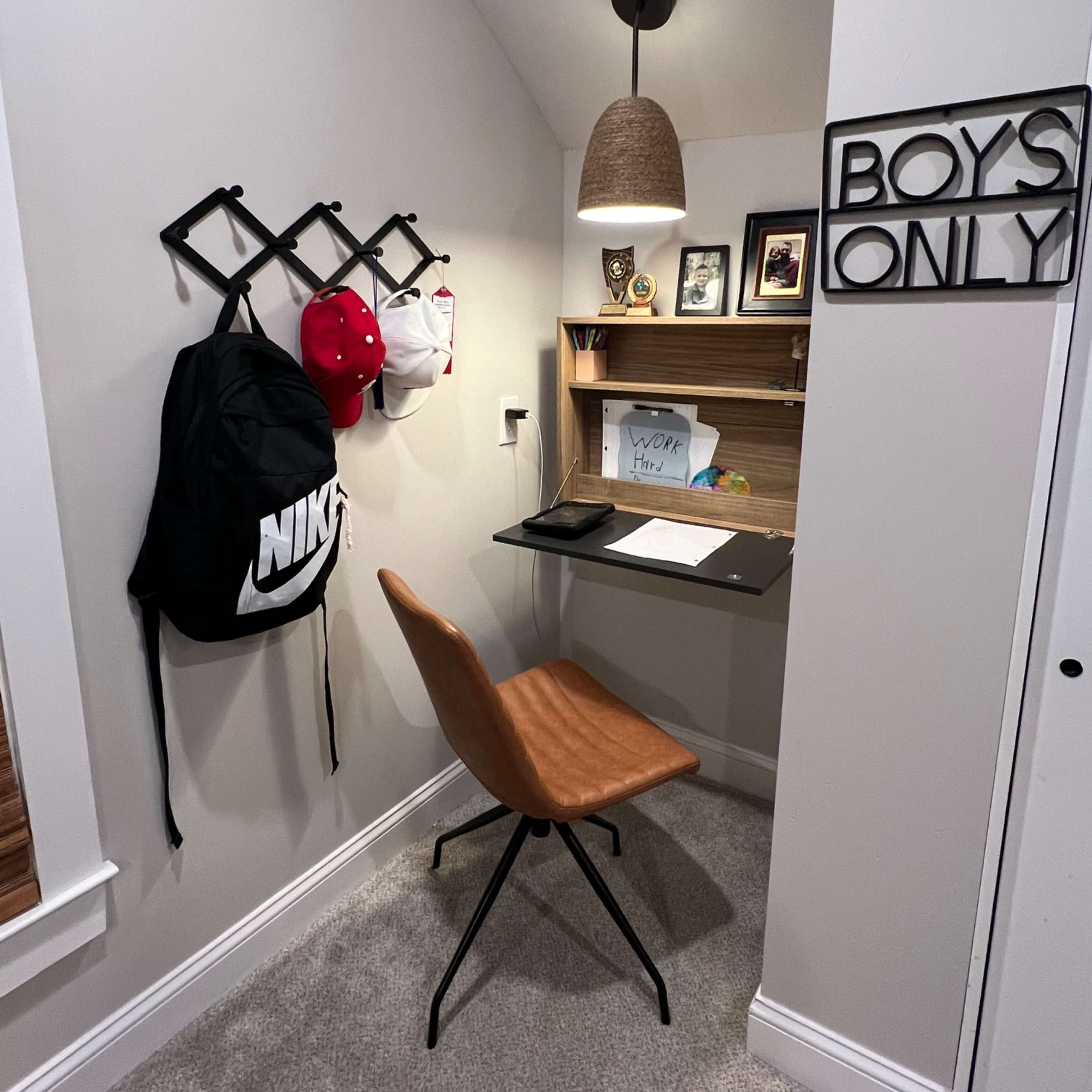 A compact workspace features a wall-mounted desk, a brown chair, backpacks hung on a rack, and a sign that reads "BOYS ONLY."