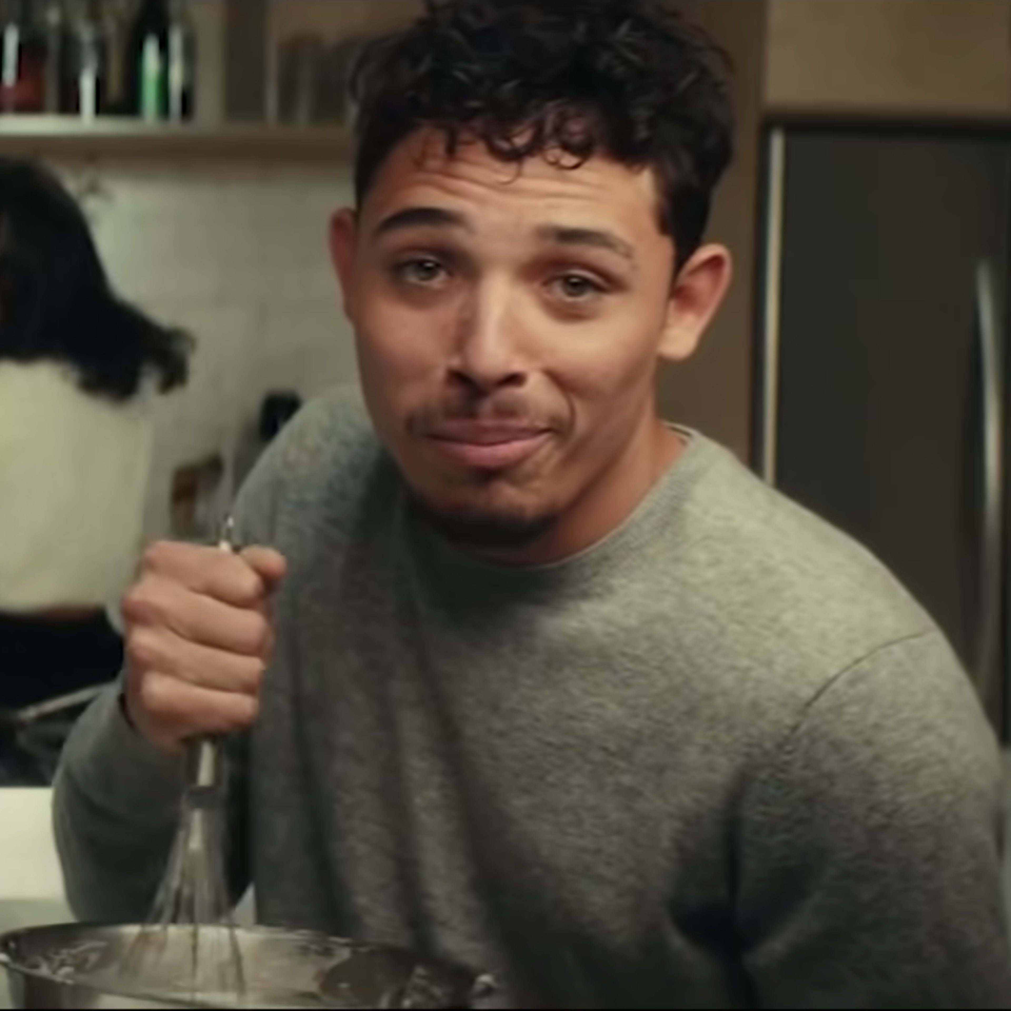 A man stands at a kitchen counter mixing ingredients while another person is visible in the background cooking.