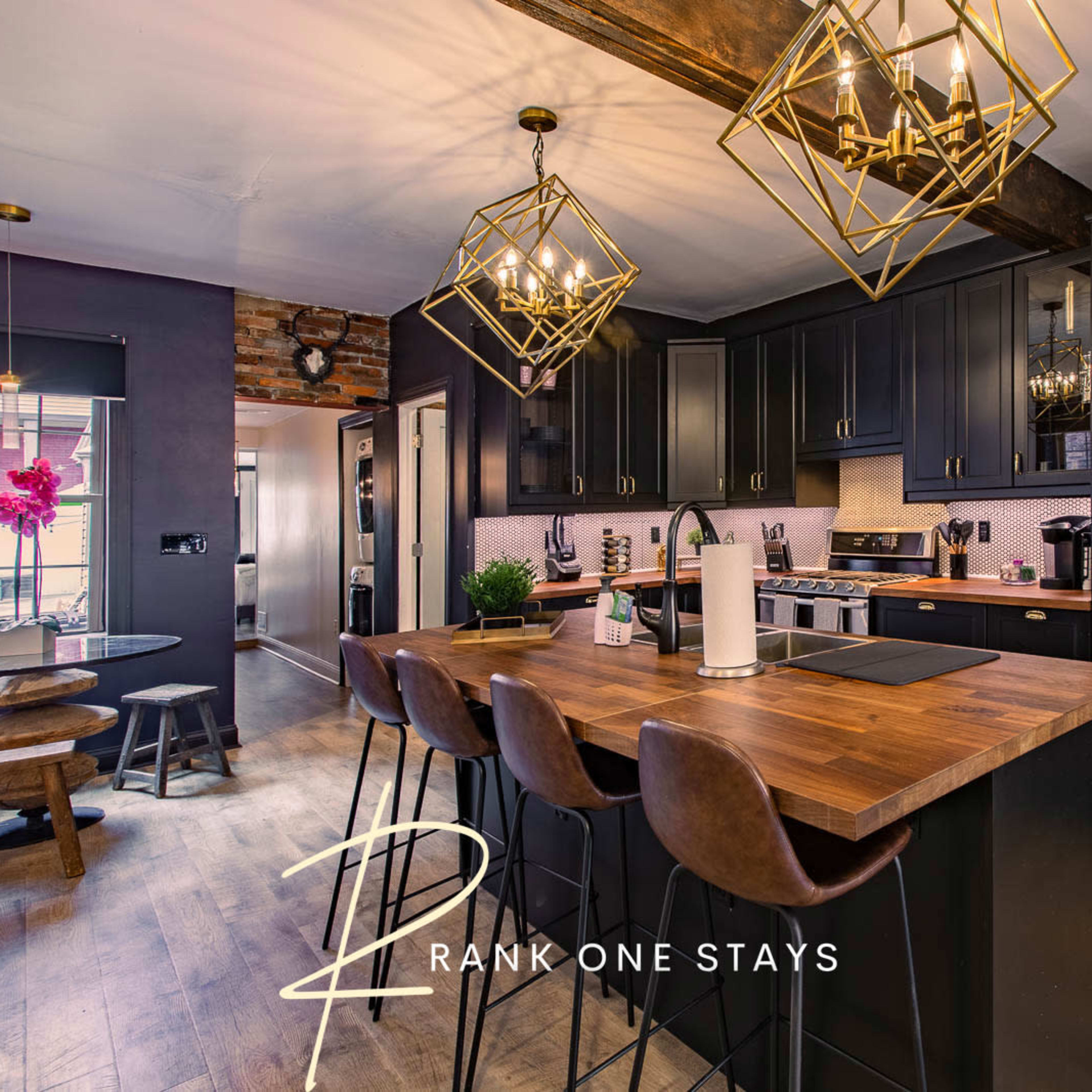 A modern kitchen with dark cabinetry, a wooden countertop, and gold pendant lights above a bar seating area.
