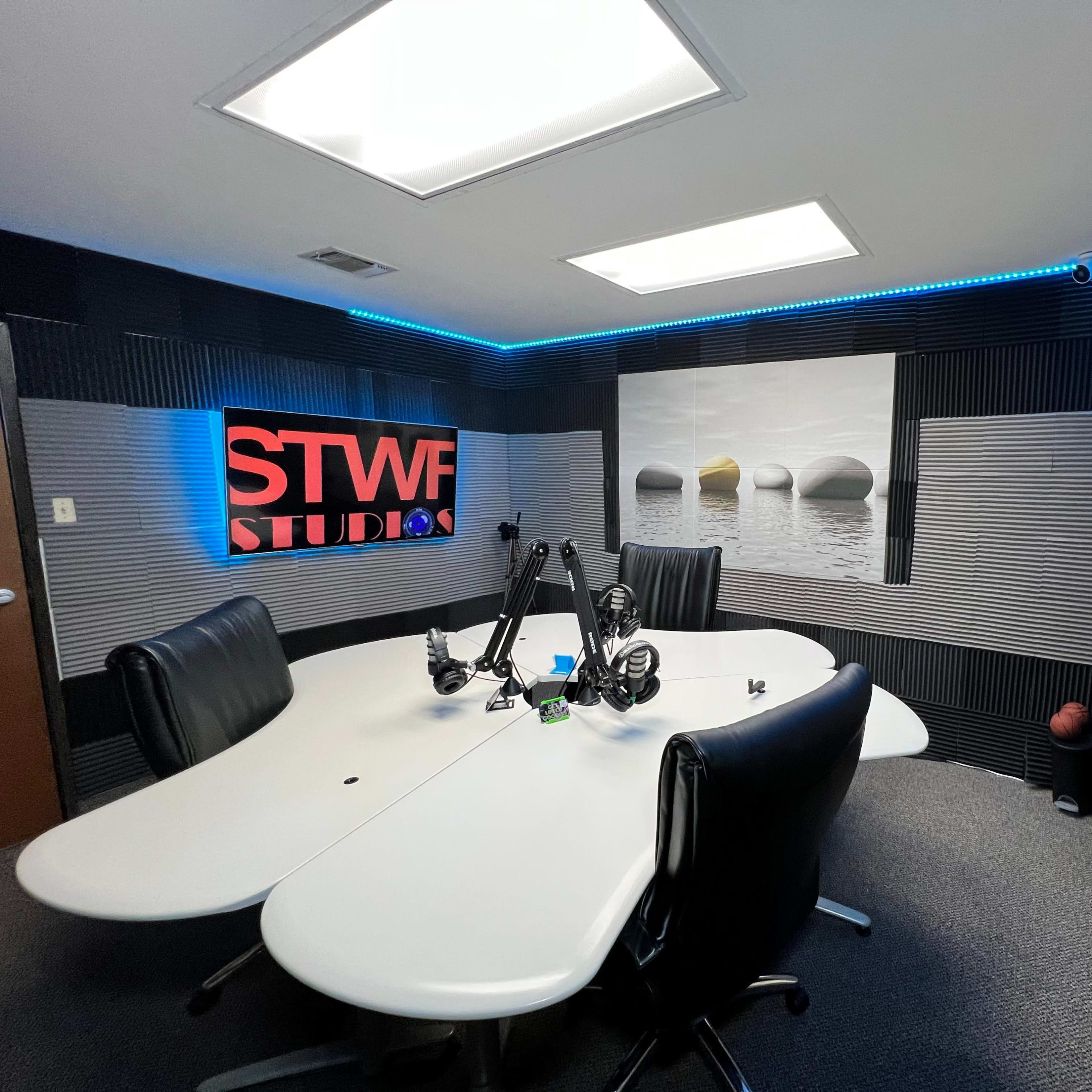 A modern recording studio featuring a large oval table surrounded by black chairs, with soundproofing panels on the walls and blue LED lights.
