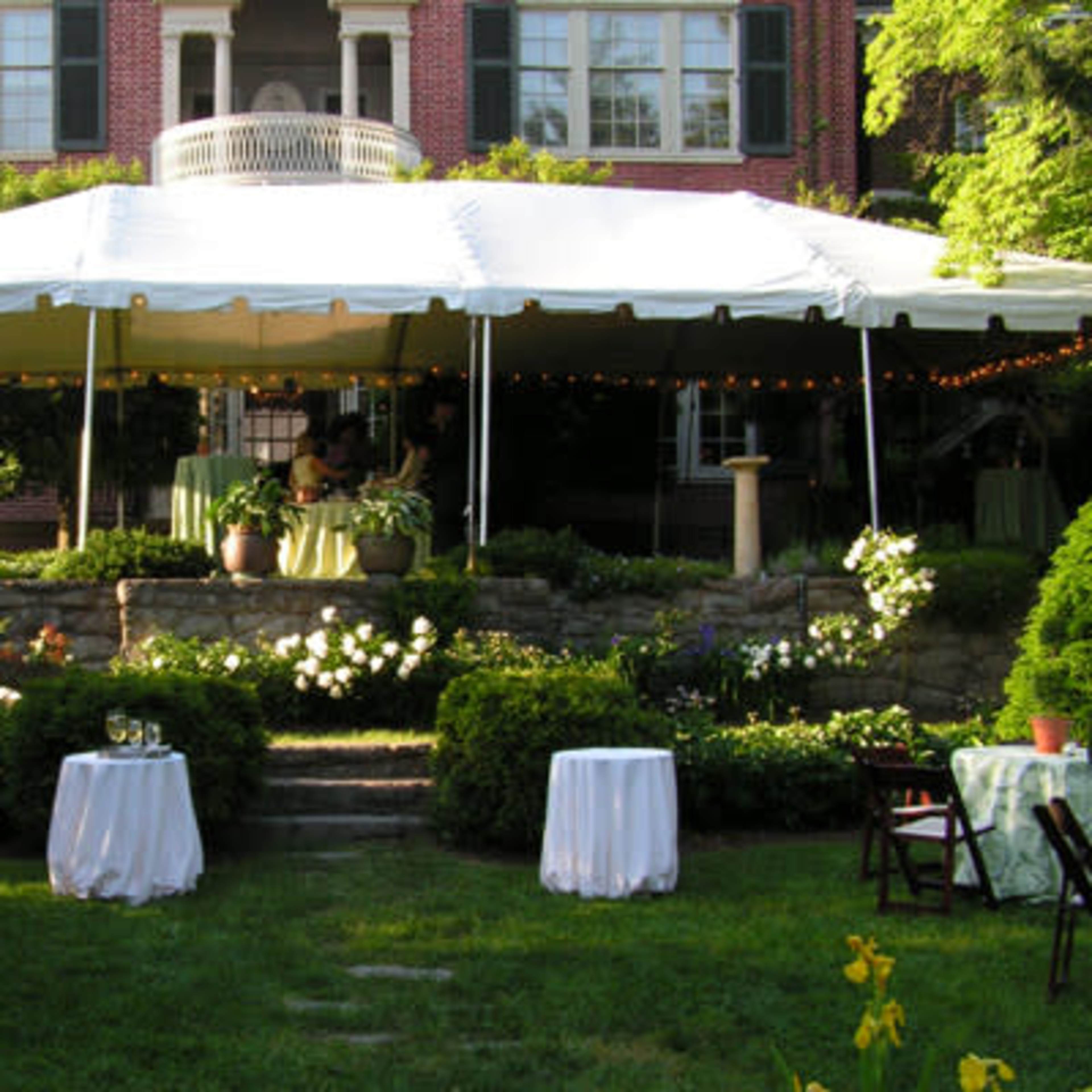 A large tent is set up in a garden area in front of a brick house, with tables and chairs arranged around it.