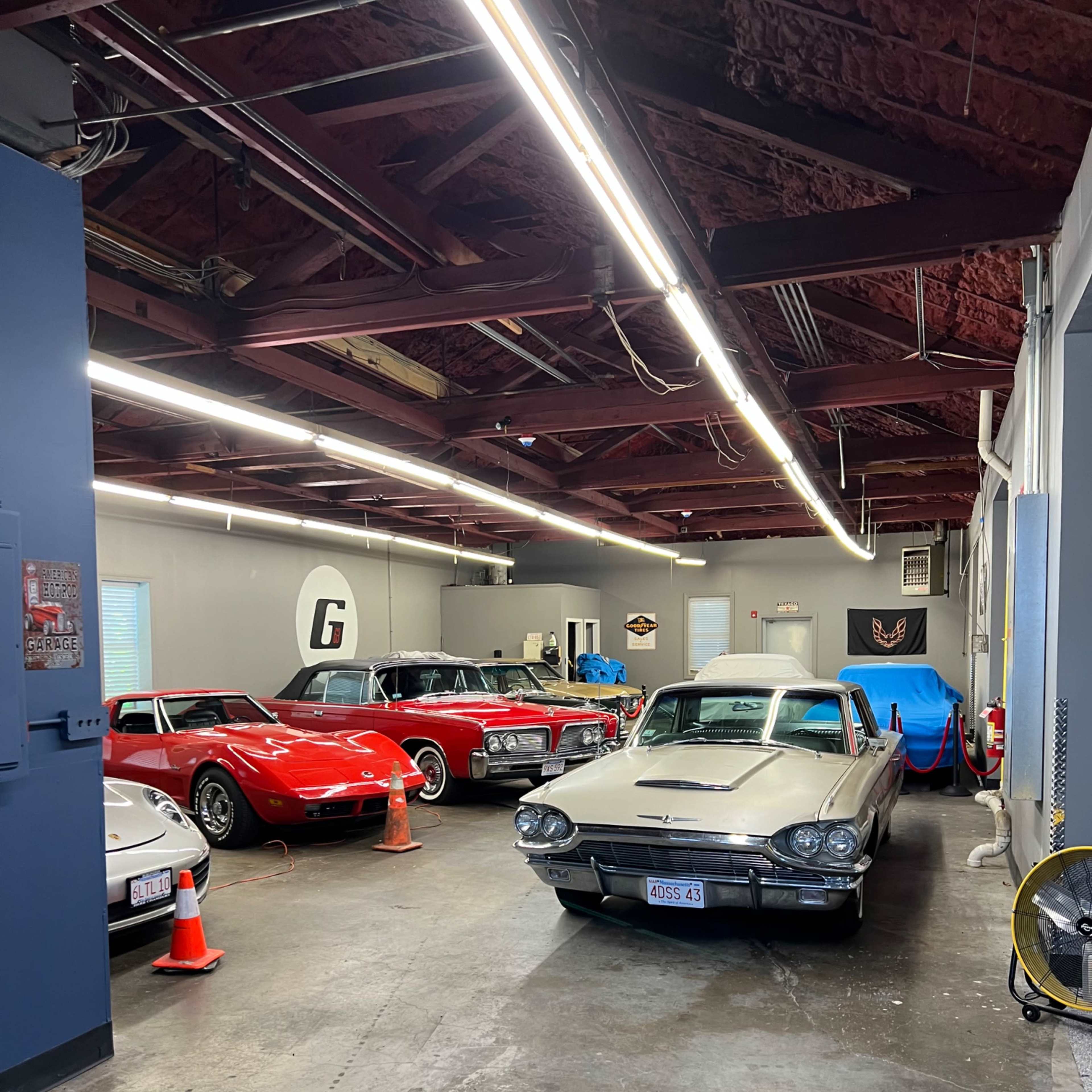 A spacious garage with several classic cars, including a silver Thunderbird and a red Corvette, parked under bright overhead lights.