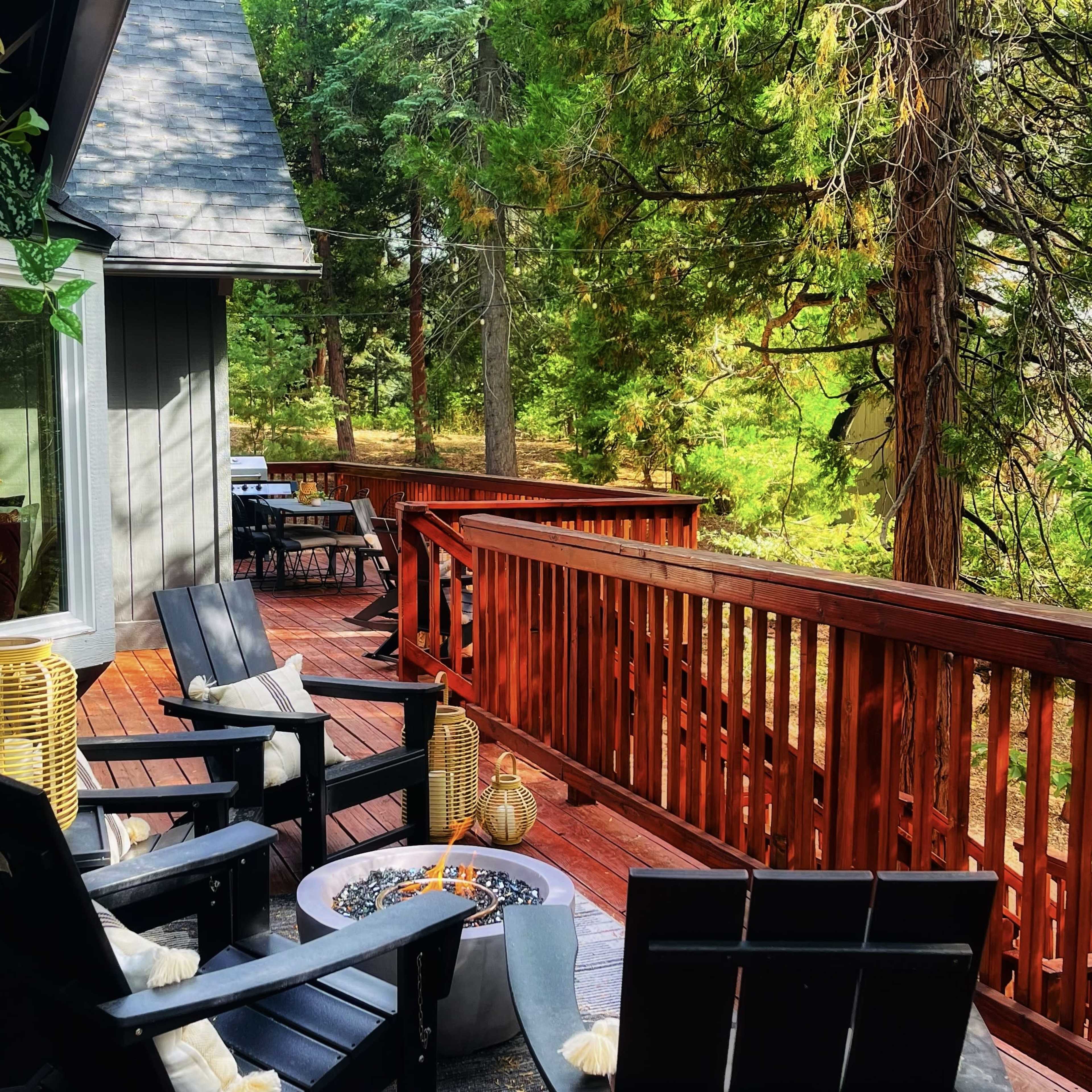A wooden deck features black lounge chairs surrounding a fire pit, with a view of green trees and outdoor seating in the background.