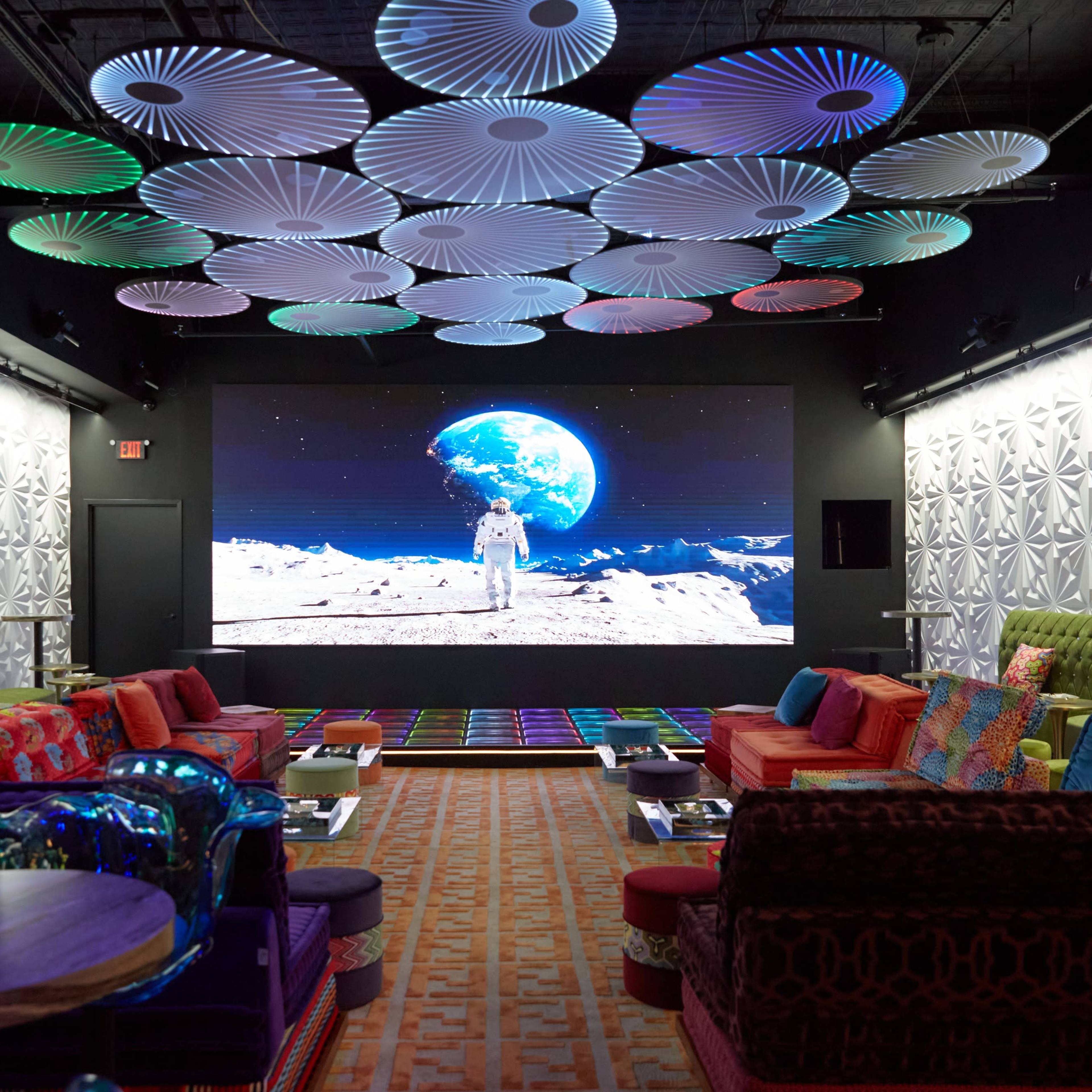 A spacious lounge features colorful seating and a large screen displaying an astronaut on the moon, surrounded by decorative, illuminated ceiling fixtures.