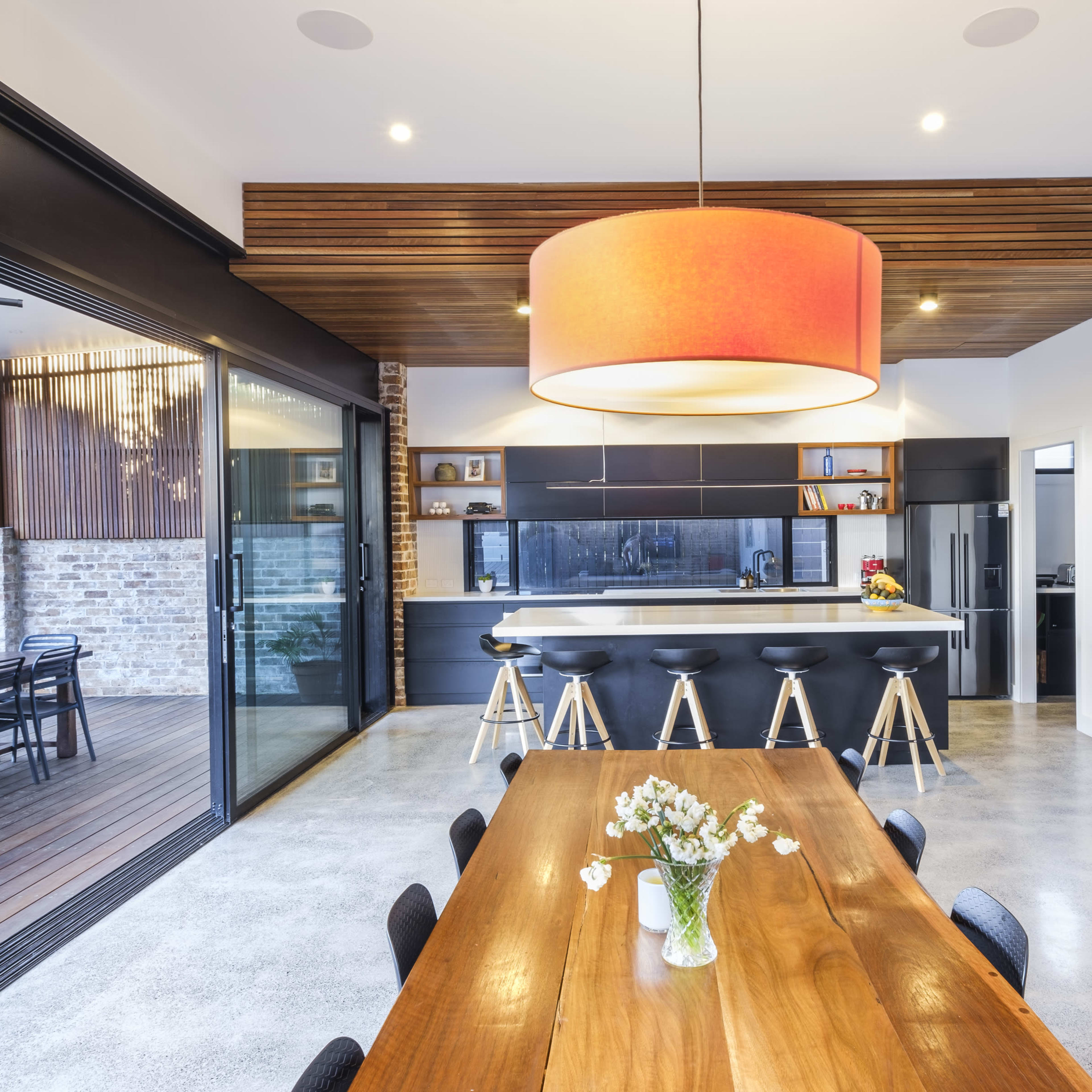 A modern kitchen and dining area featuring a large wooden table, black bar stools at a kitchen island, and large glass doors leading to an outdoor patio.