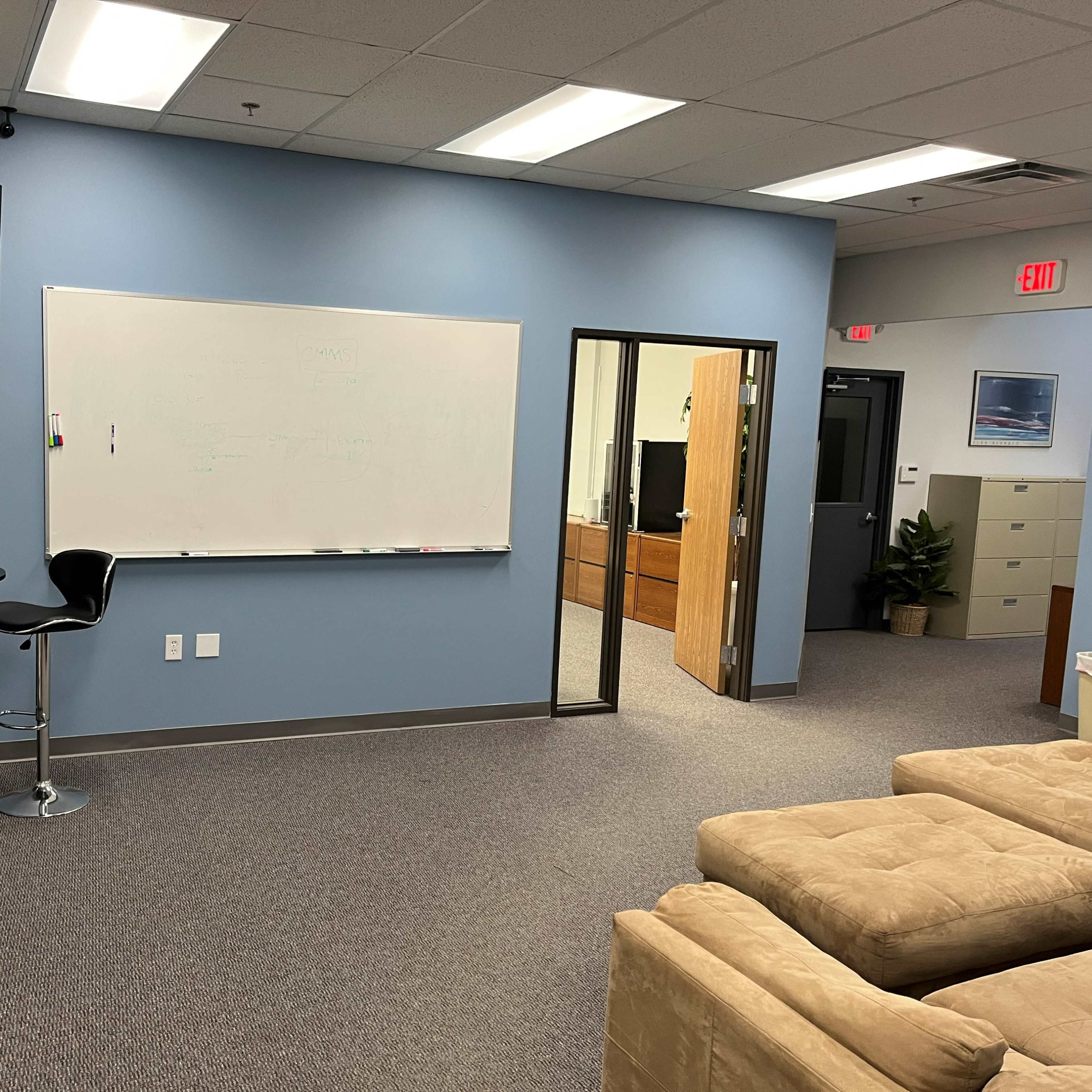 An office space with a seating area, a whiteboard, a bar table with a stool, a television mounted on the wall, and a doorway leading to another room.