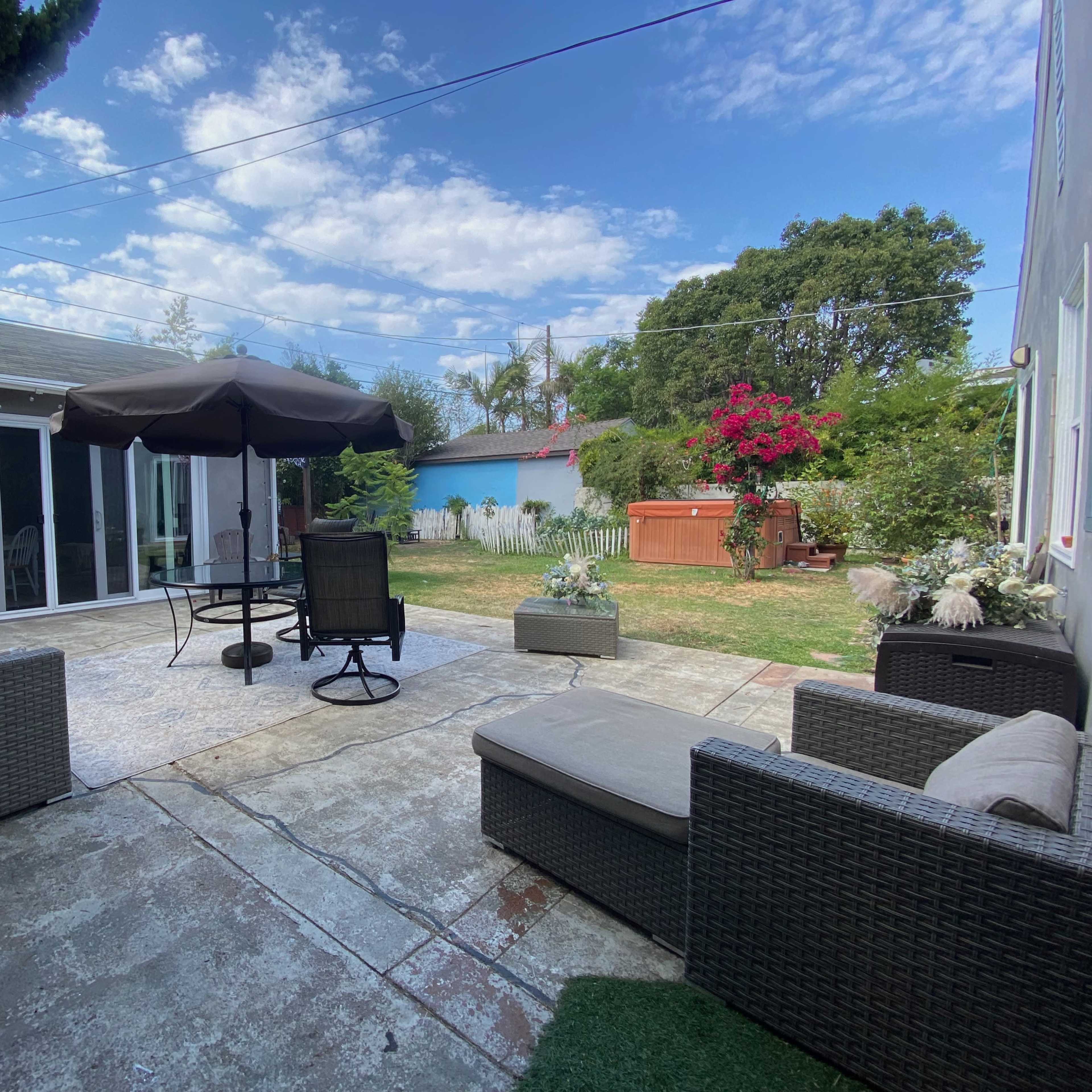 A patio area furnished with seating, a table with an umbrella, and a lawn surrounded by greenery and flowering plants.