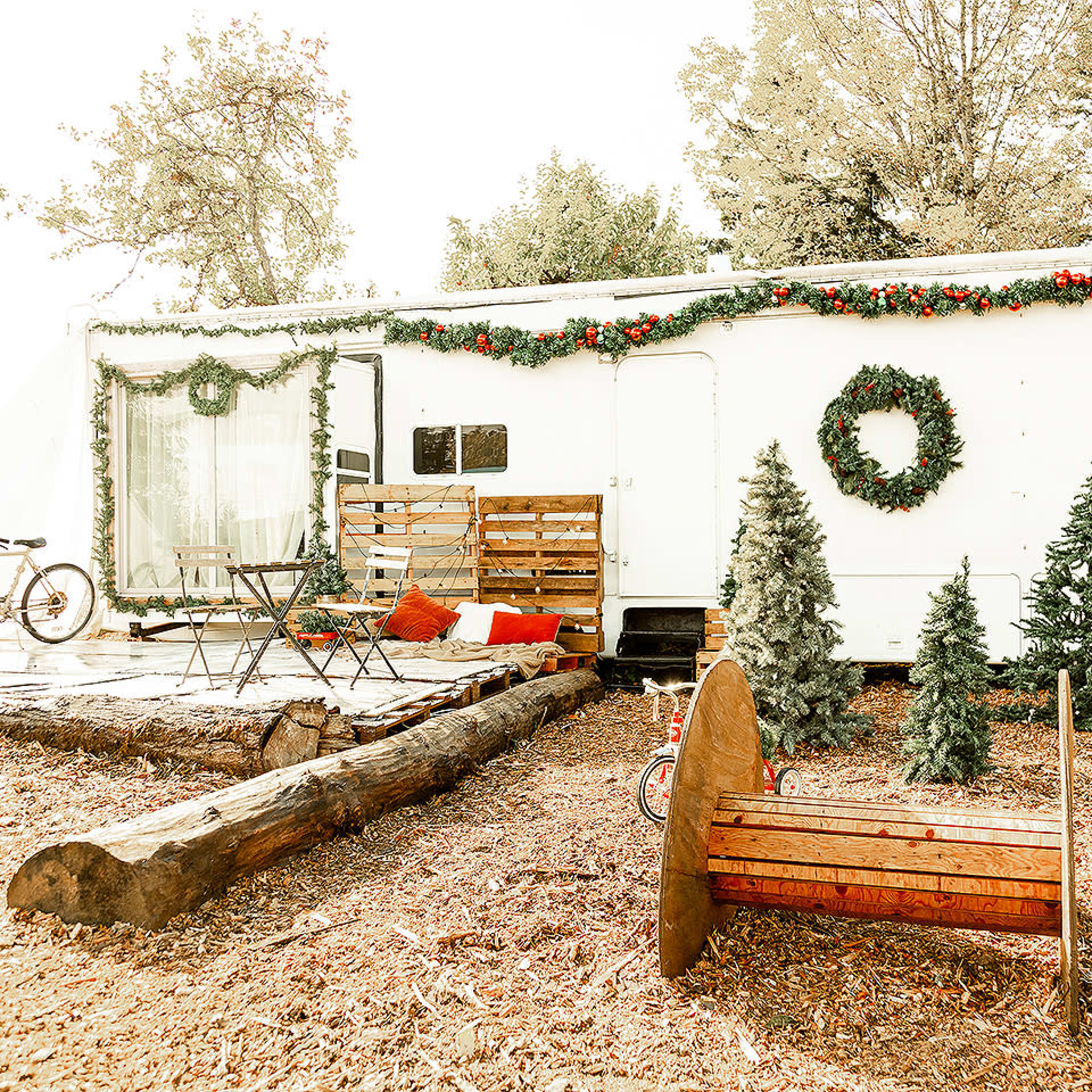 A white camper trailer is decorated with greenery and surrounded by artificial Christmas trees, a wooden seating area, and a bicycle.