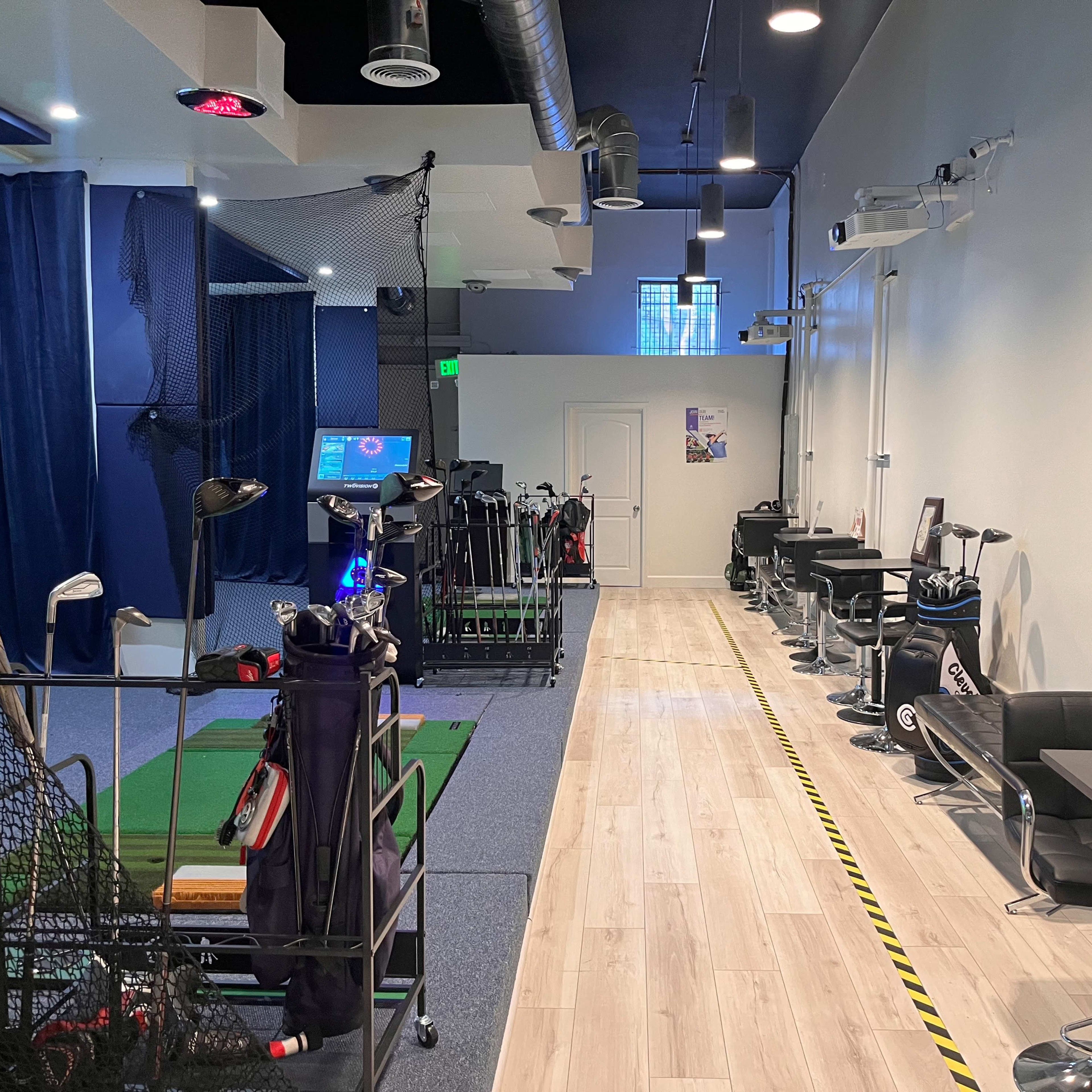 An indoor golf simulation facility with a carpeted area for practice on one side and tables and chairs along the opposite wall.