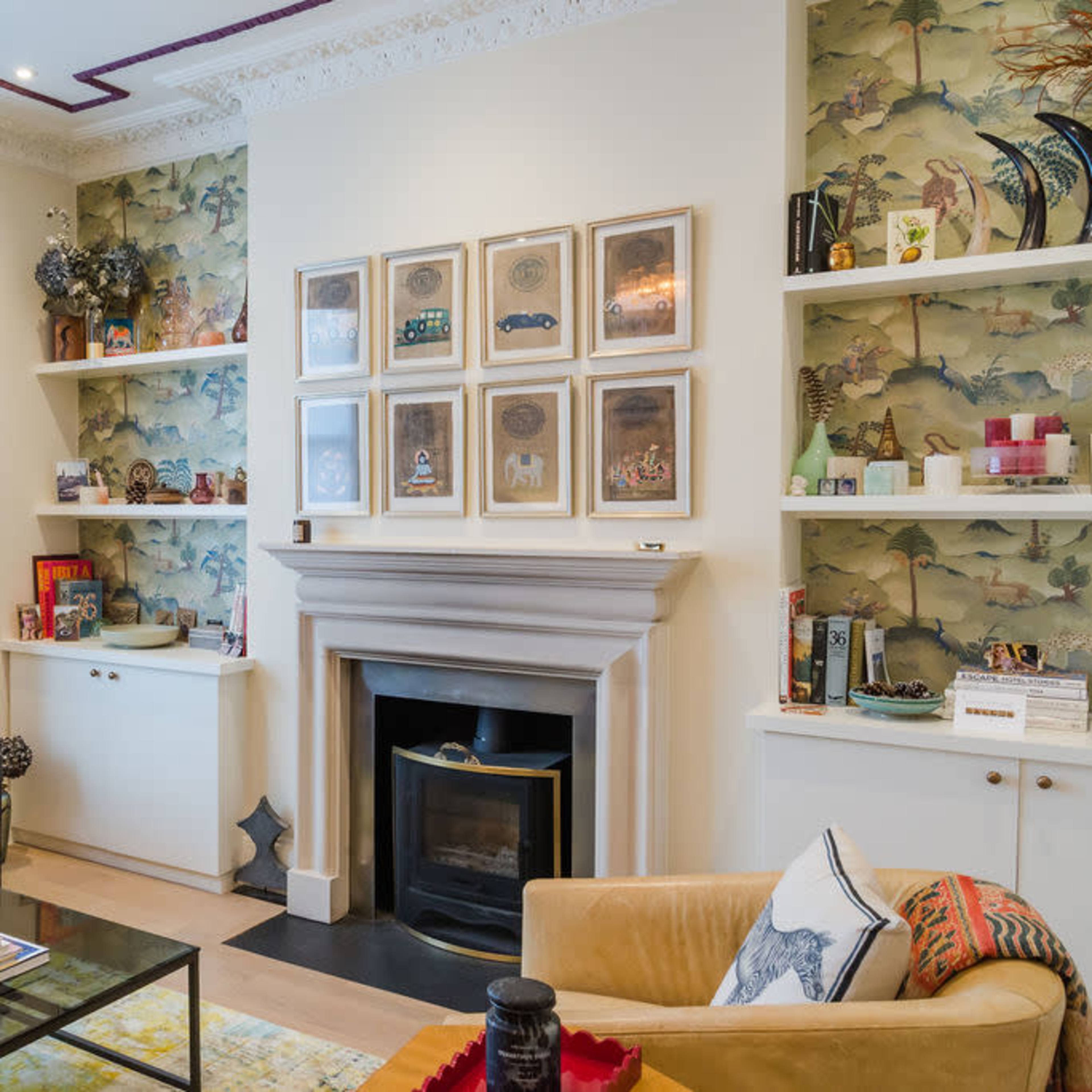 A living room featuring a fireplace, bookshelves with decorative items, and layered seating arrangements, with patterned wallpaper and a chandelier overhead.