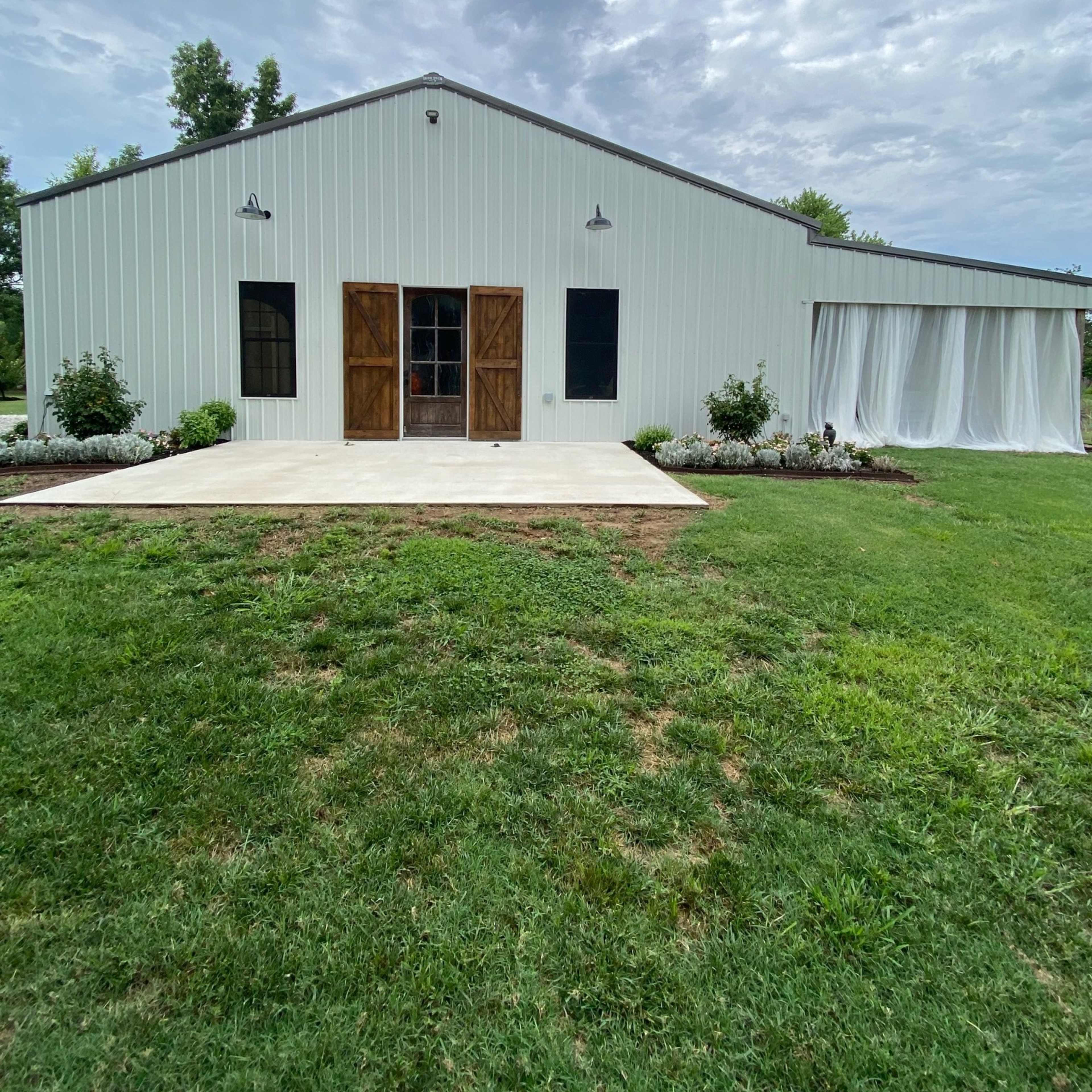 A large white metal building with dual wooden doors and windows stands on a grassy area, featuring a covered patio with landscaping in front.