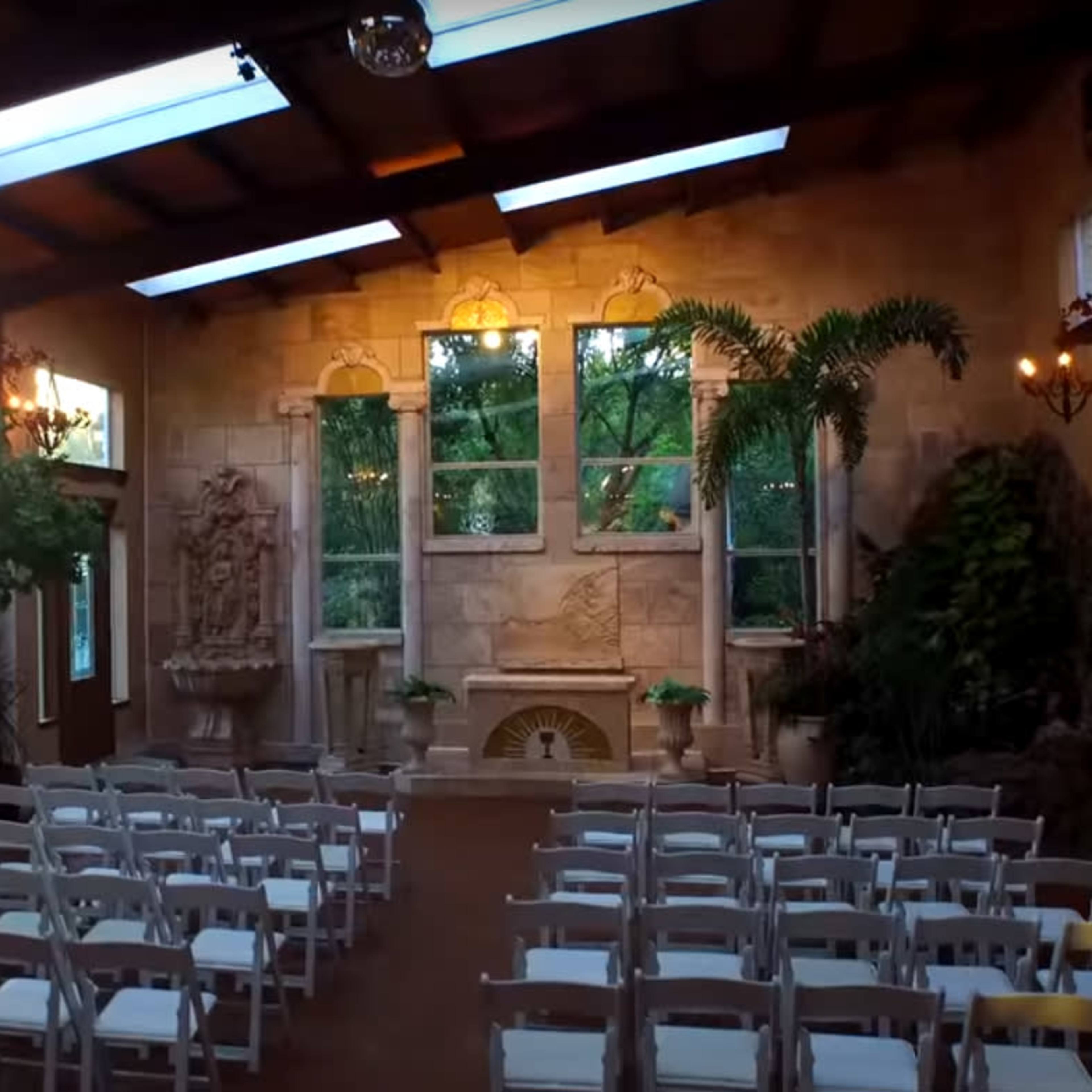 A wedding ceremony setup with rows of white chairs arranged in front of a decorative stone backdrop featuring large windows and greenery.