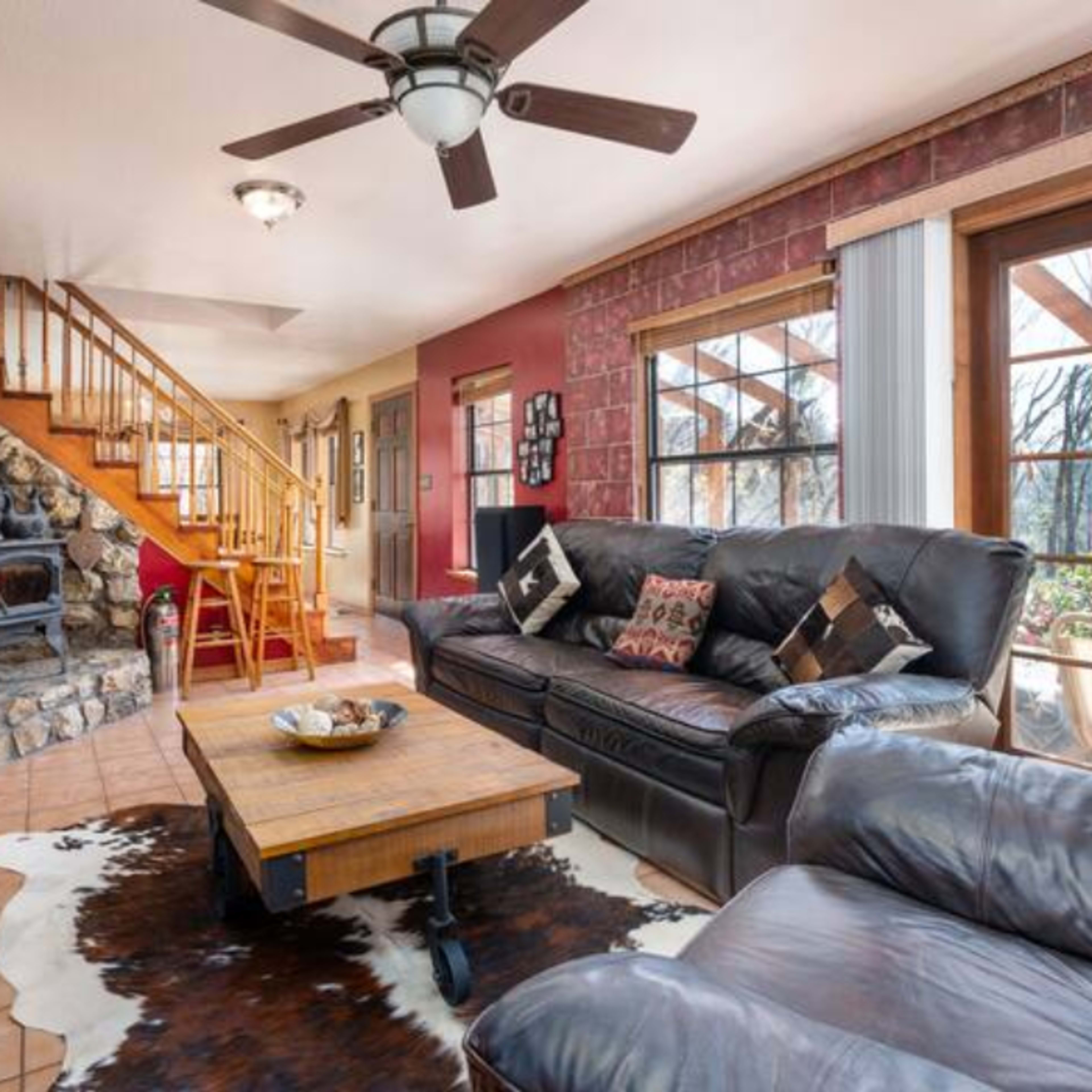 A living room with a stone fireplace, a wooden staircase, a coffee table, and large windows leading to a deck.