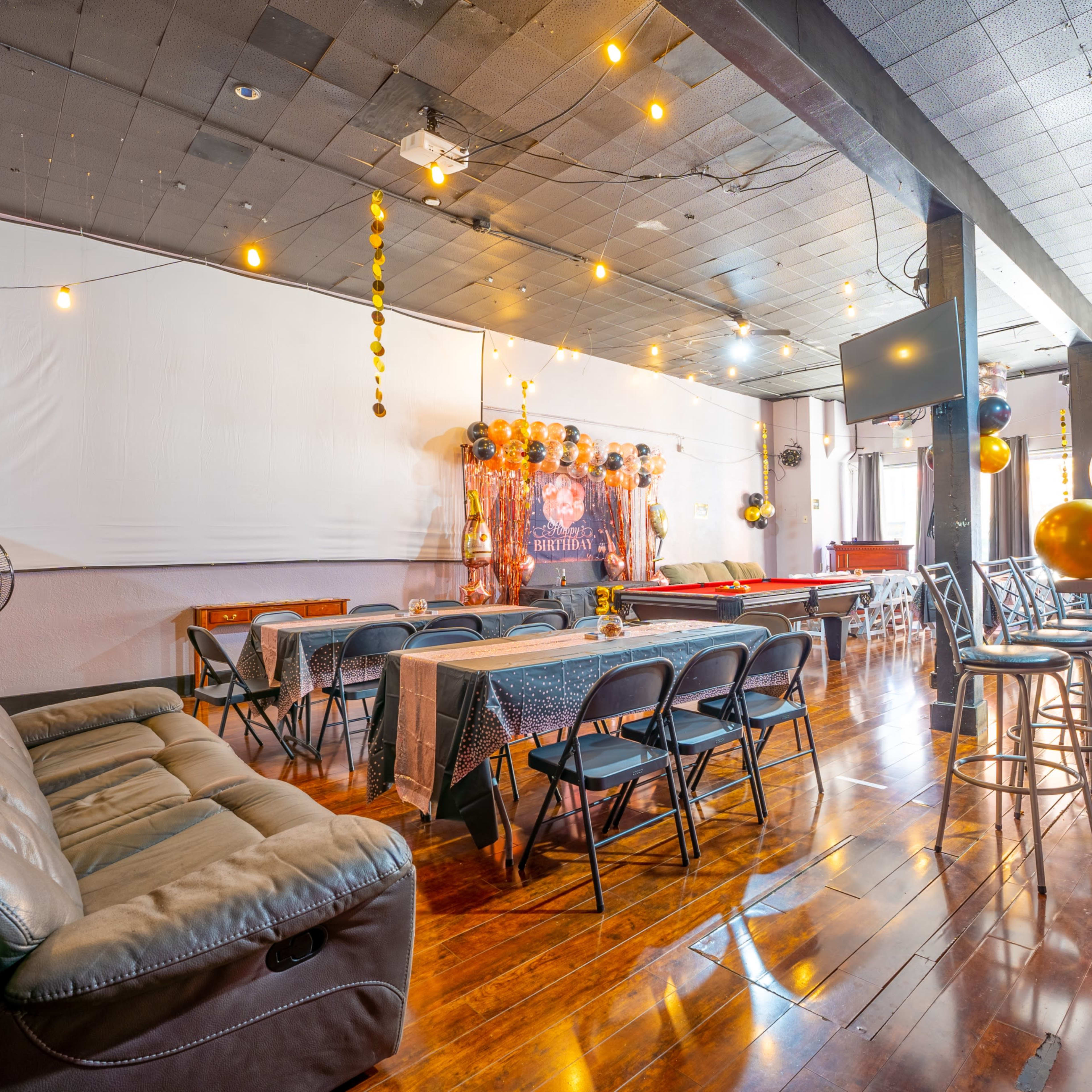 A spacious room with wooden flooring, featuring a seating area with a leather couch, tables covered with cloths, and a projection screen on the wall, decorated for an event.