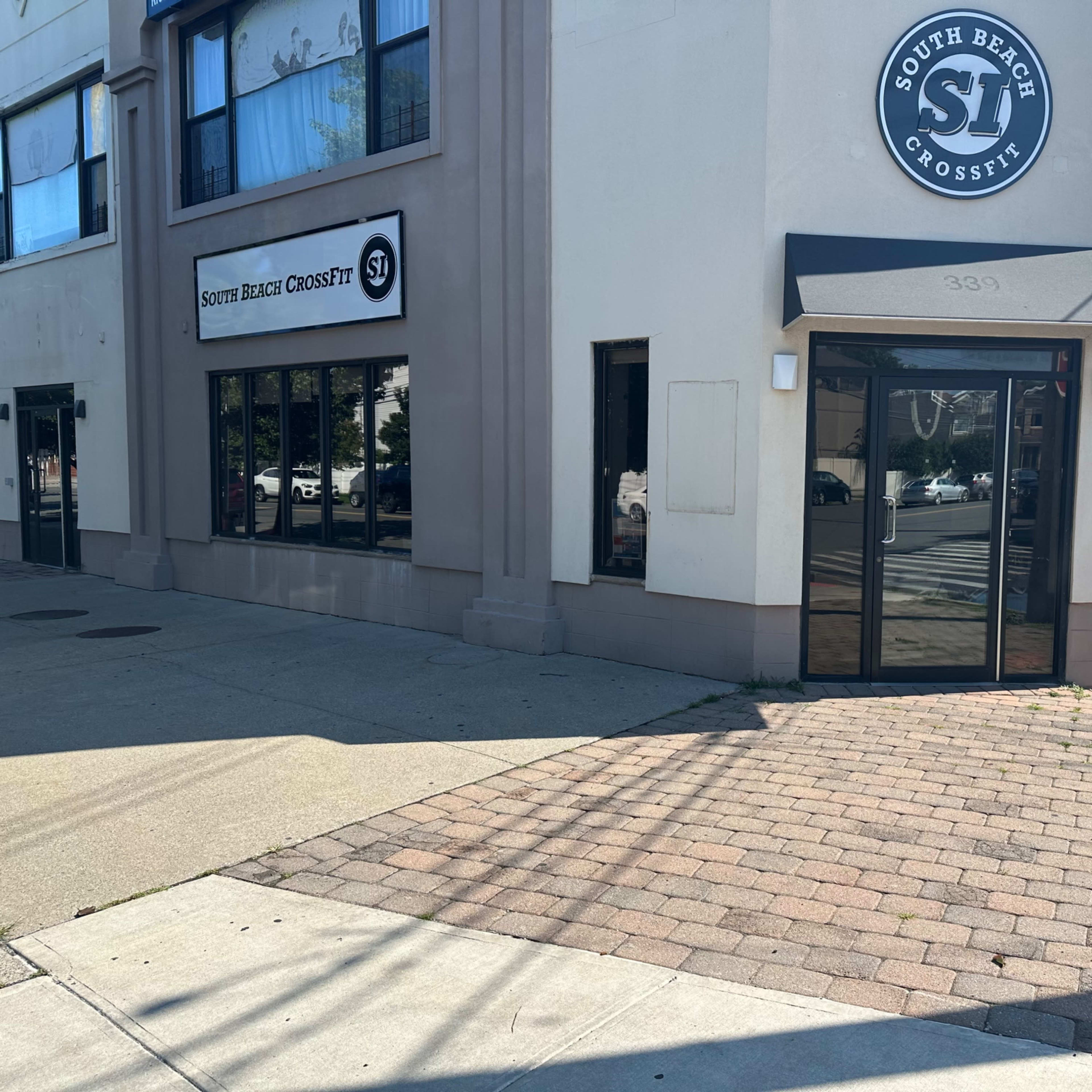 The entrance of South Beach CrossFit, a fitness facility located on the corner of a building, with a paved sidewalk and parked cars nearby.