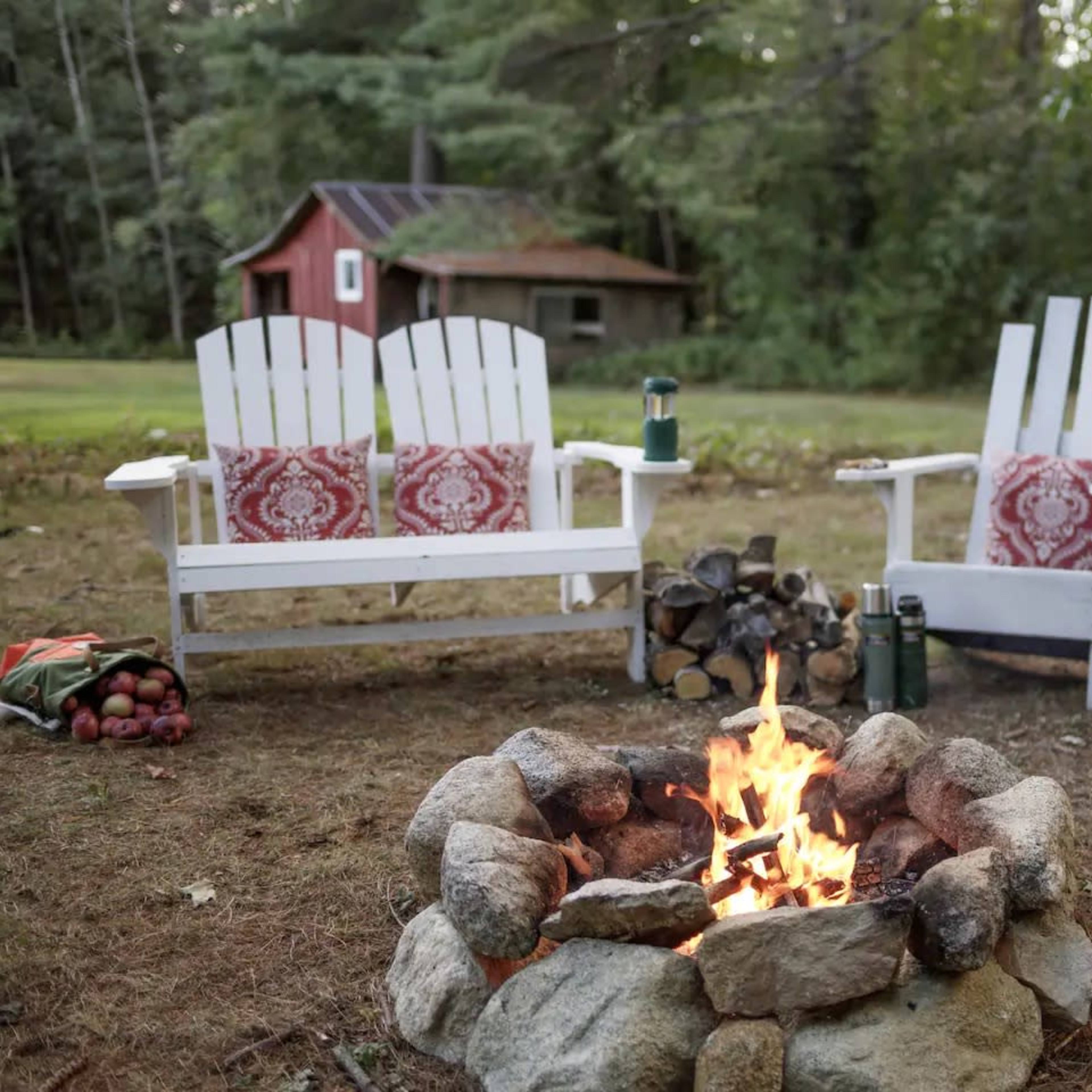 Two white Adirondack chairs with red cushions sit beside a stone fire pit, surrounded by a grassy area and a rustic shed in the background.