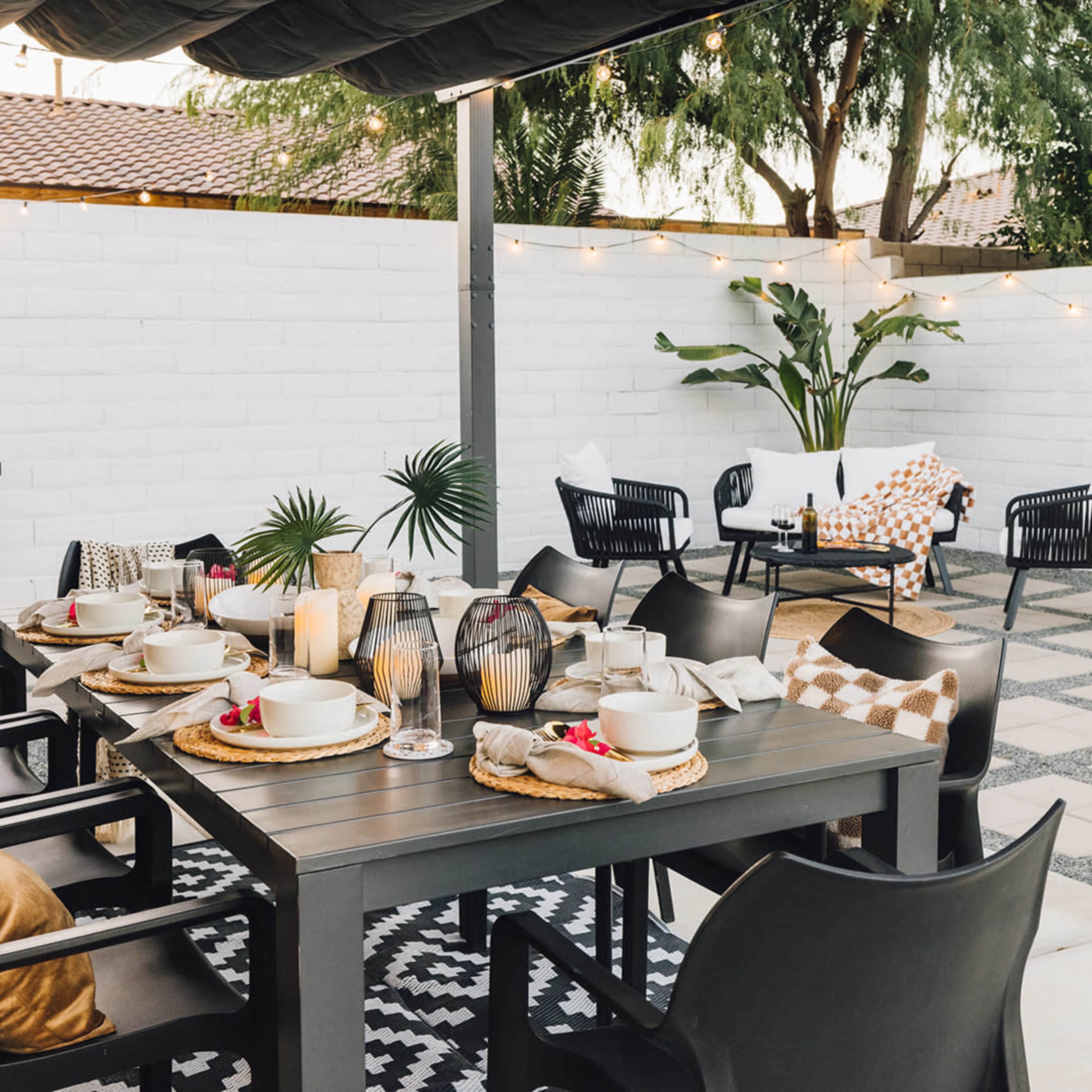 The outdoor dining area features a black table set with dishes and candles, surrounded by chairs and a barbecue grill, with string lights and potted plants in a fenced space.