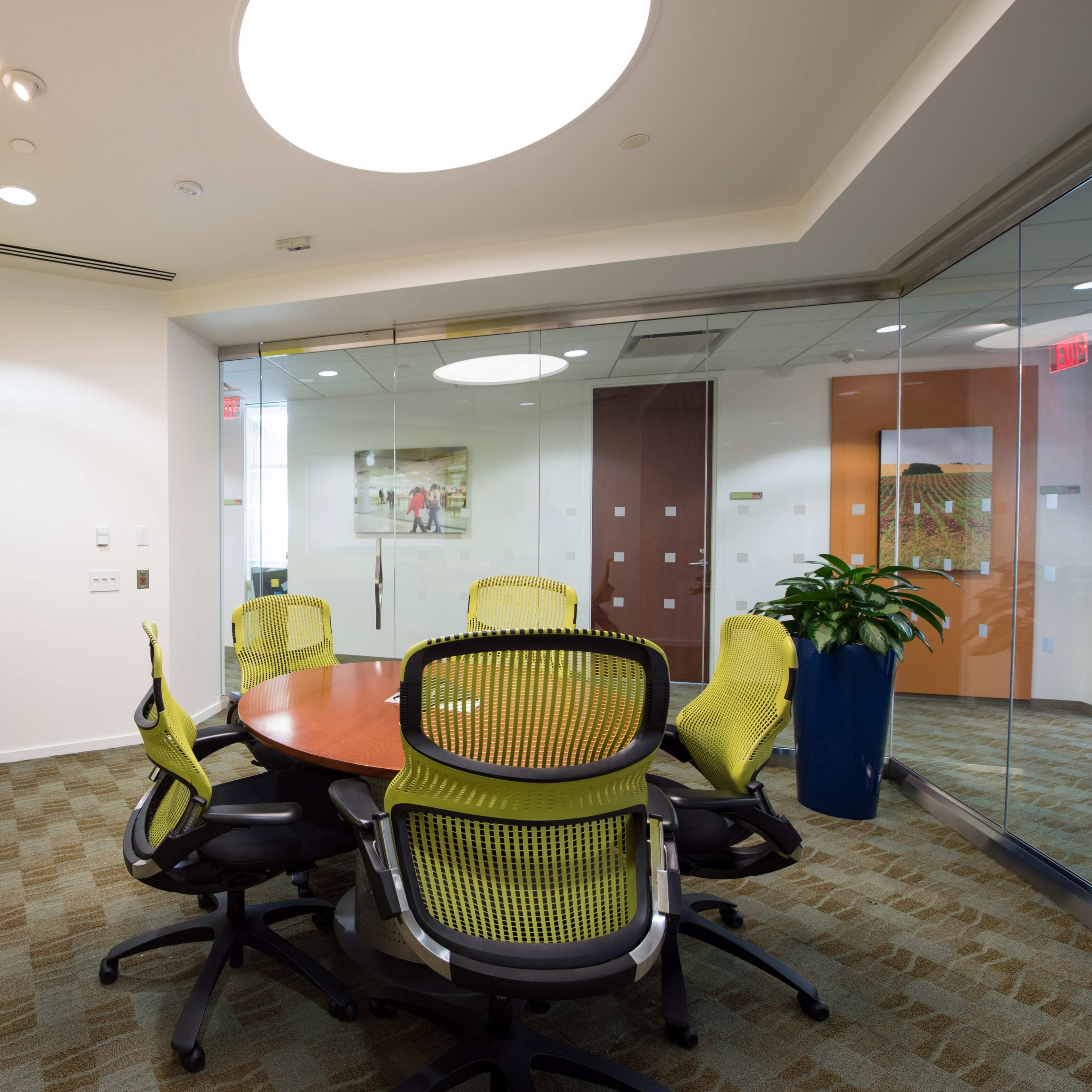 A modern conference room featuring a round table surrounded by ergonomic chairs, with a whiteboard on one wall and glass partitions.