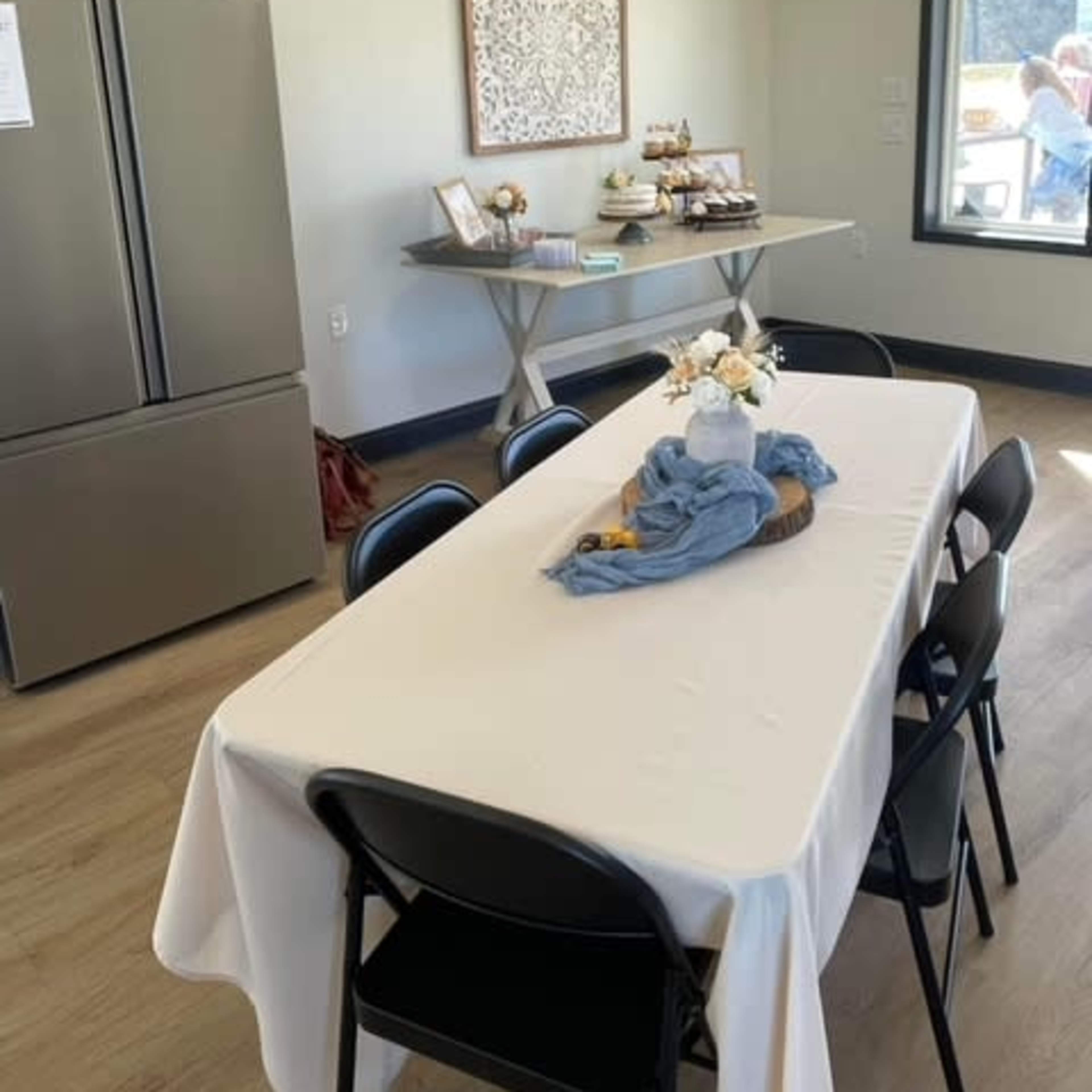 A dining area features a table covered in a white tablecloth with a centerpiece, surrounded by black chairs, alongside a gray refrigerator and a side table displaying decorative items.