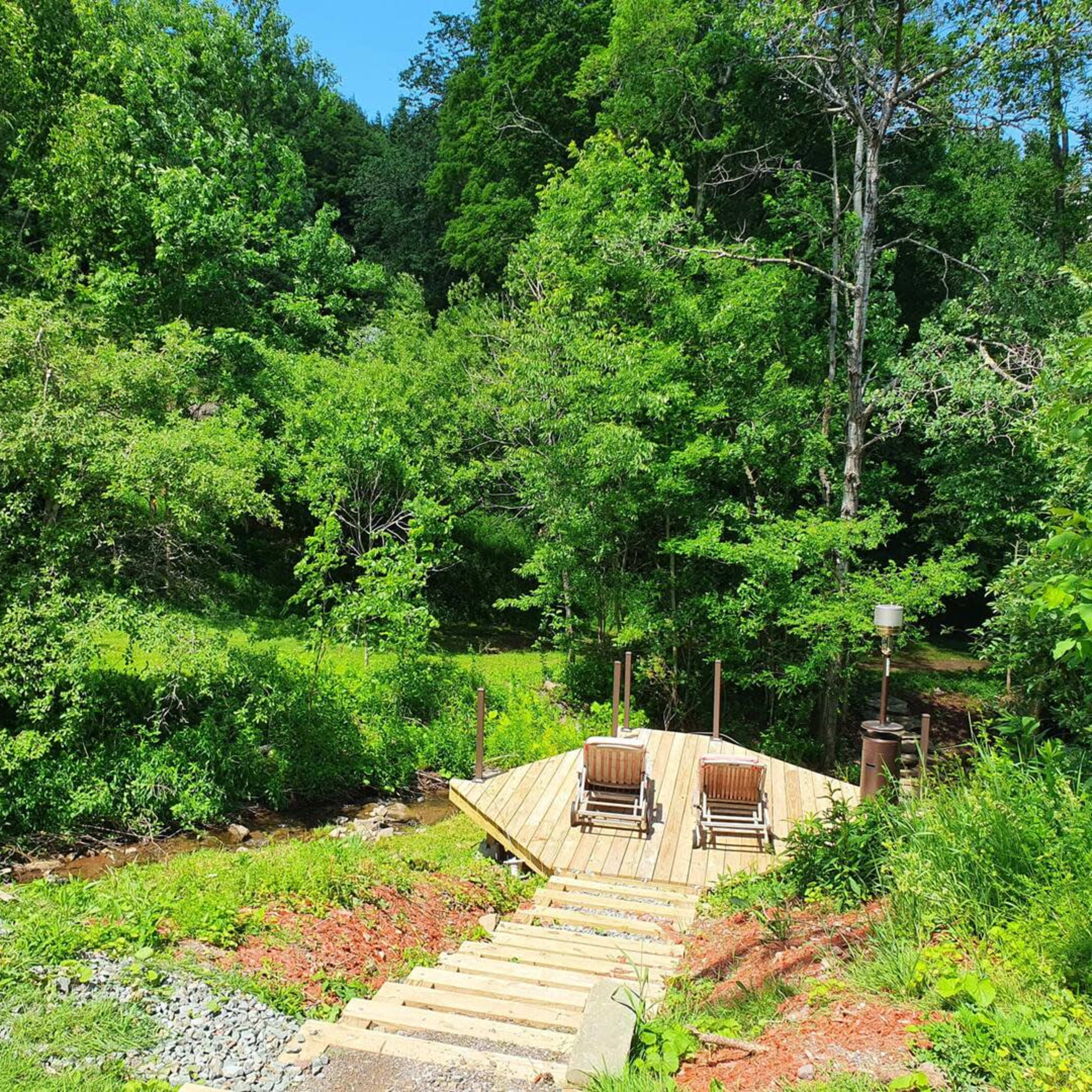 A wooden deck with two chairs overlooks a lush green forest and stream.