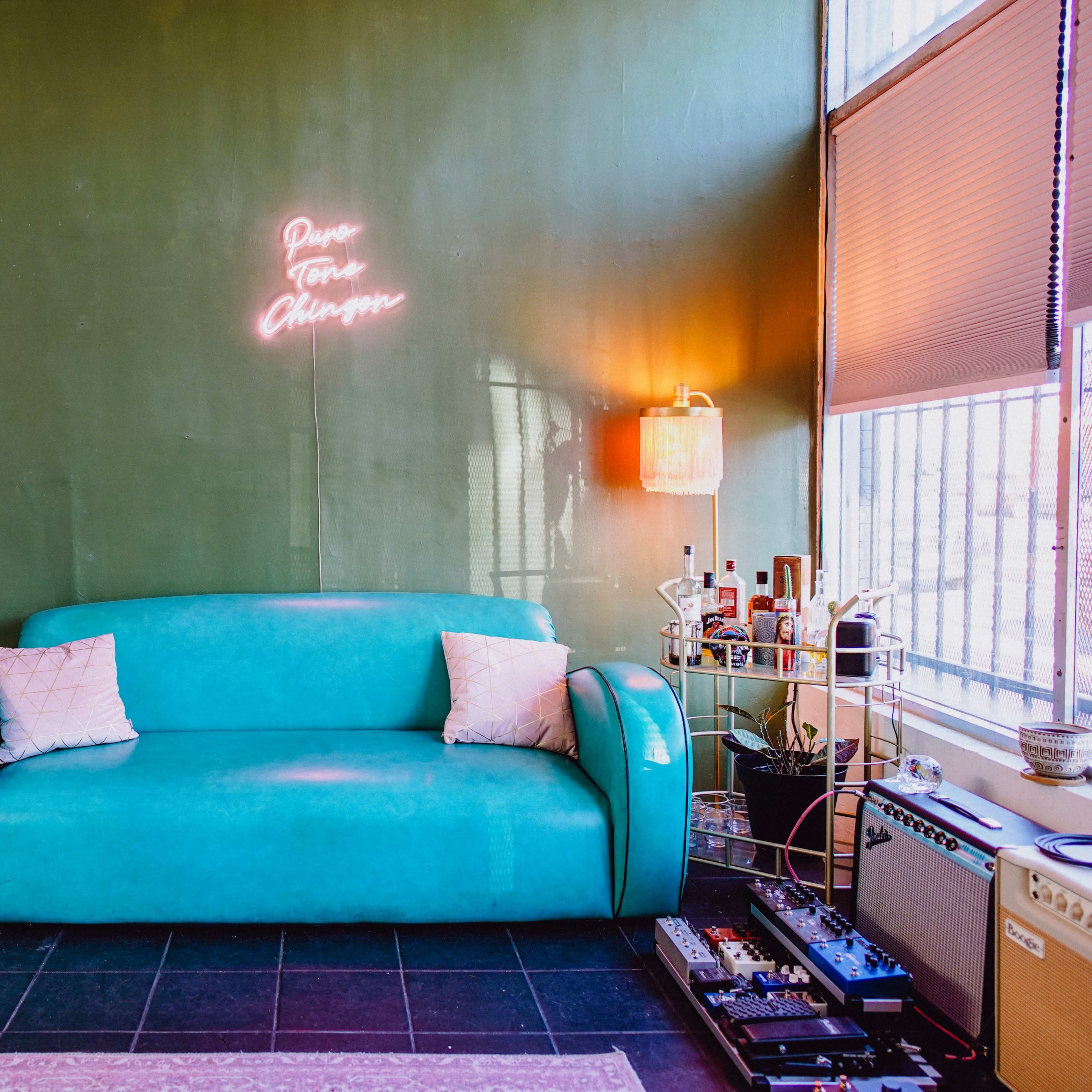 A teal sofa is positioned against a green wall with a neon sign, while nearby furniture and decor create a cozy living space.