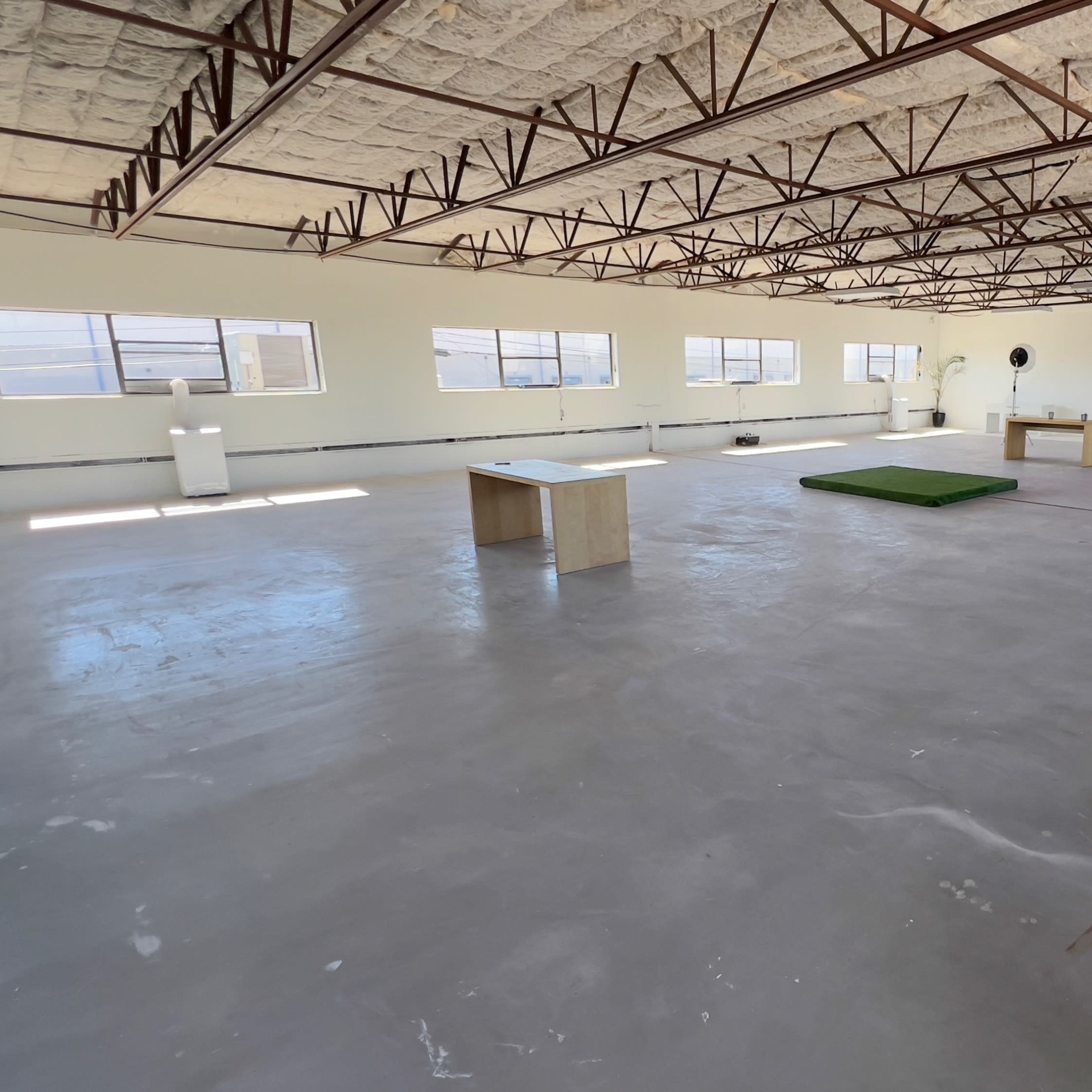 A large, empty room with a concrete floor, several windows, and minimal furnishings including a table and a green mat.