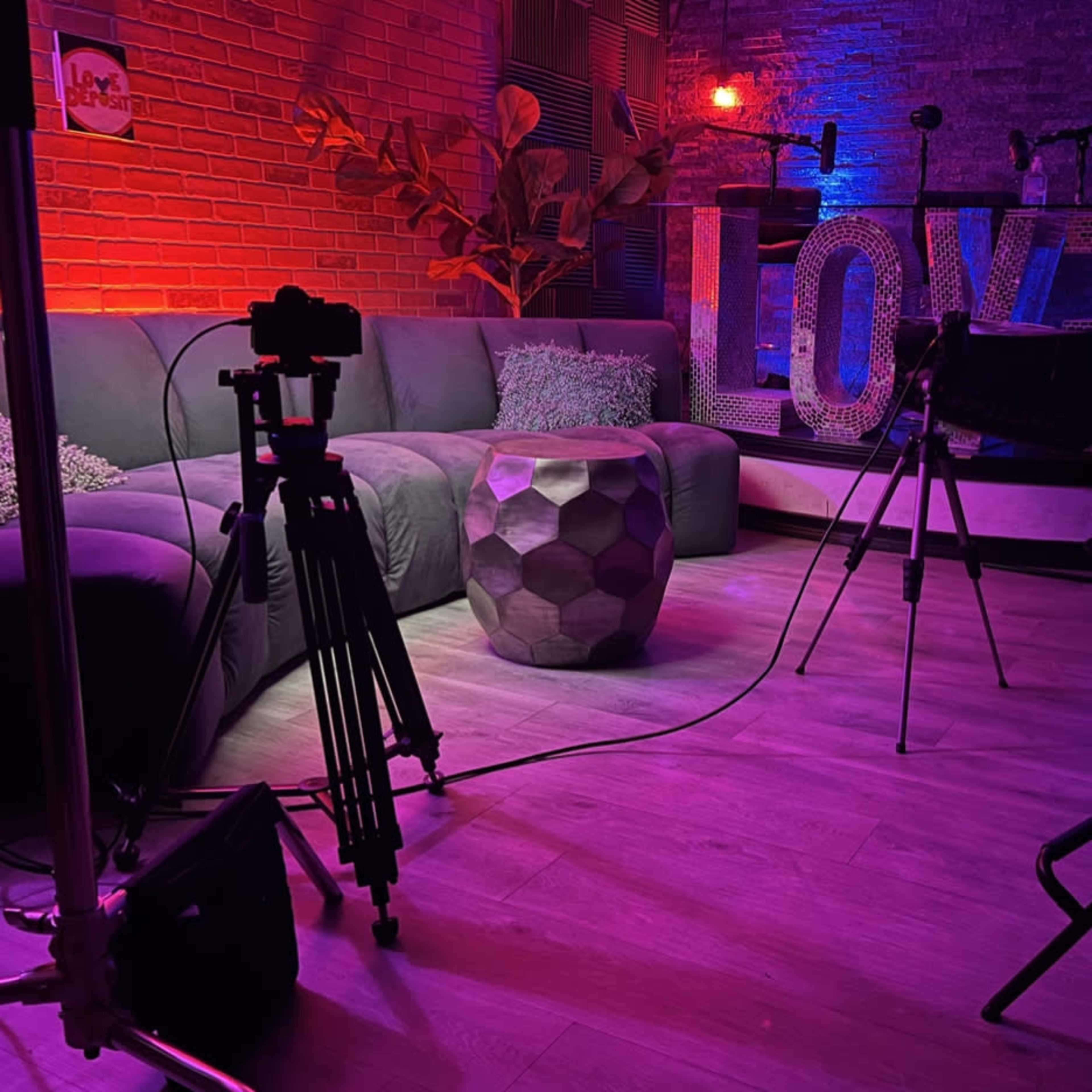 A dimly lit studio setup with a green sofa, a geometric coffee table, and a large "LOVE" sign illuminated in the background.