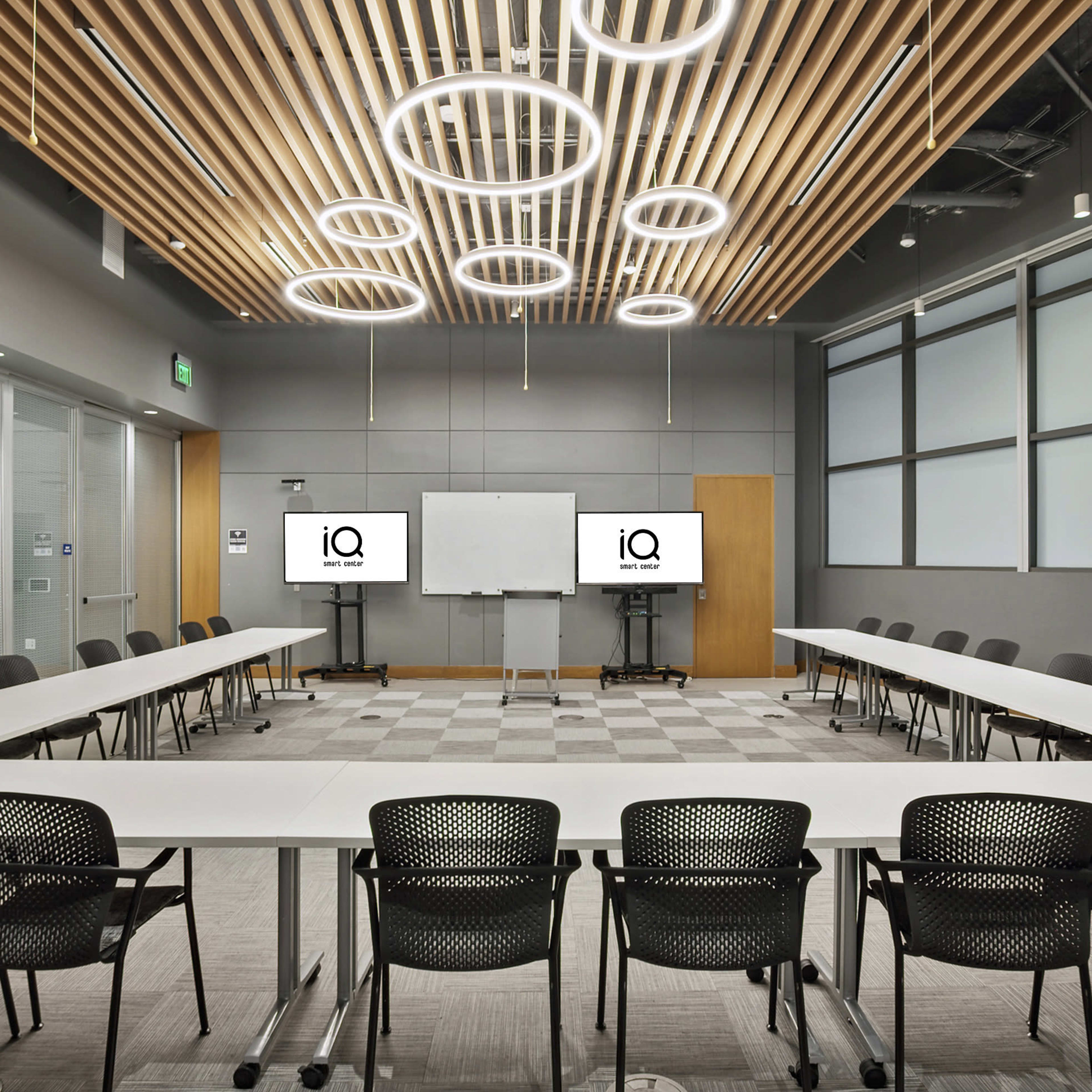 A modern conference room features a rectangular table surrounded by black chairs, with two screens displaying the letters "iQ" and a whiteboard at one end.