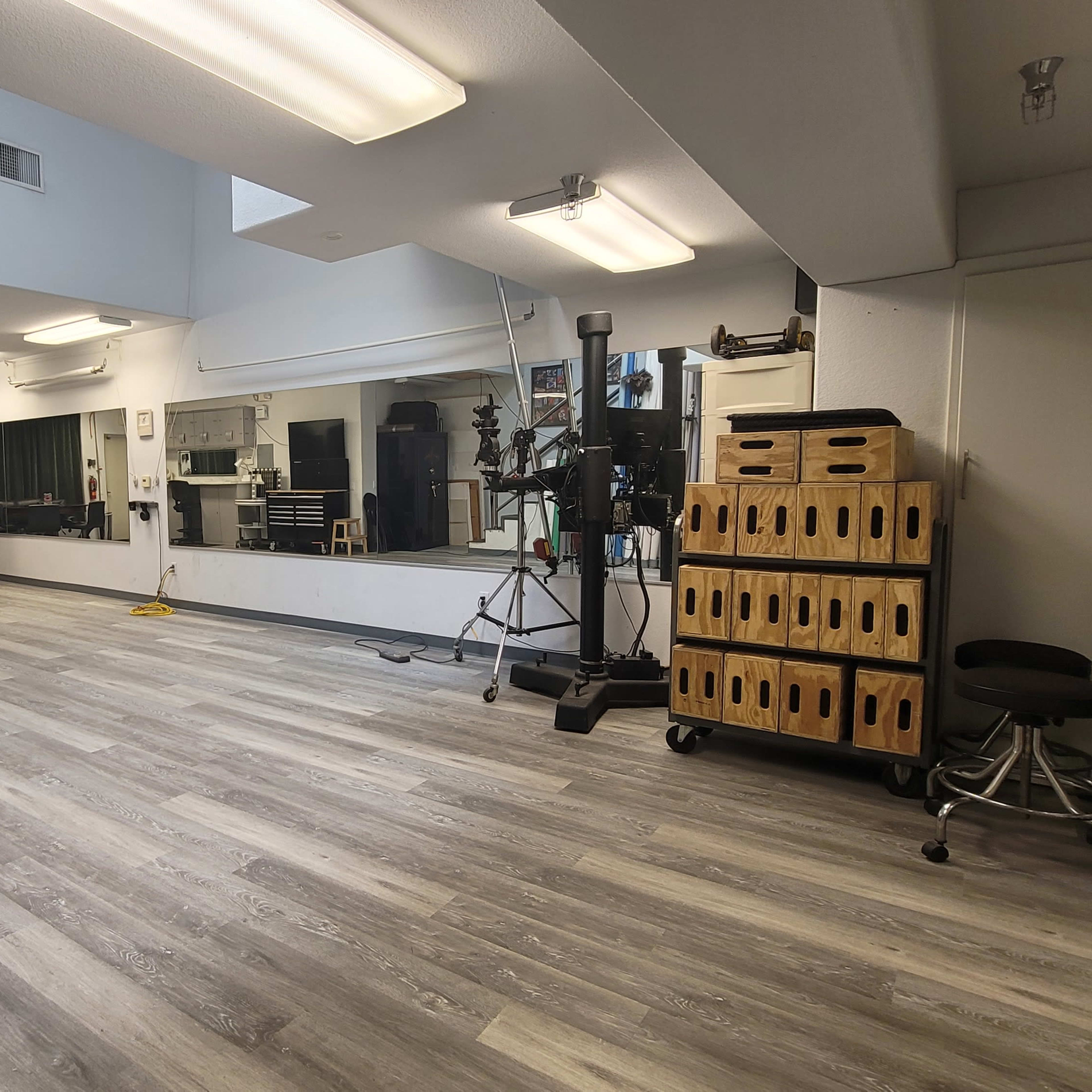 A large, empty room with gray wood flooring, equipped with mirrors, studio lighting, and storage boxes along the wall.