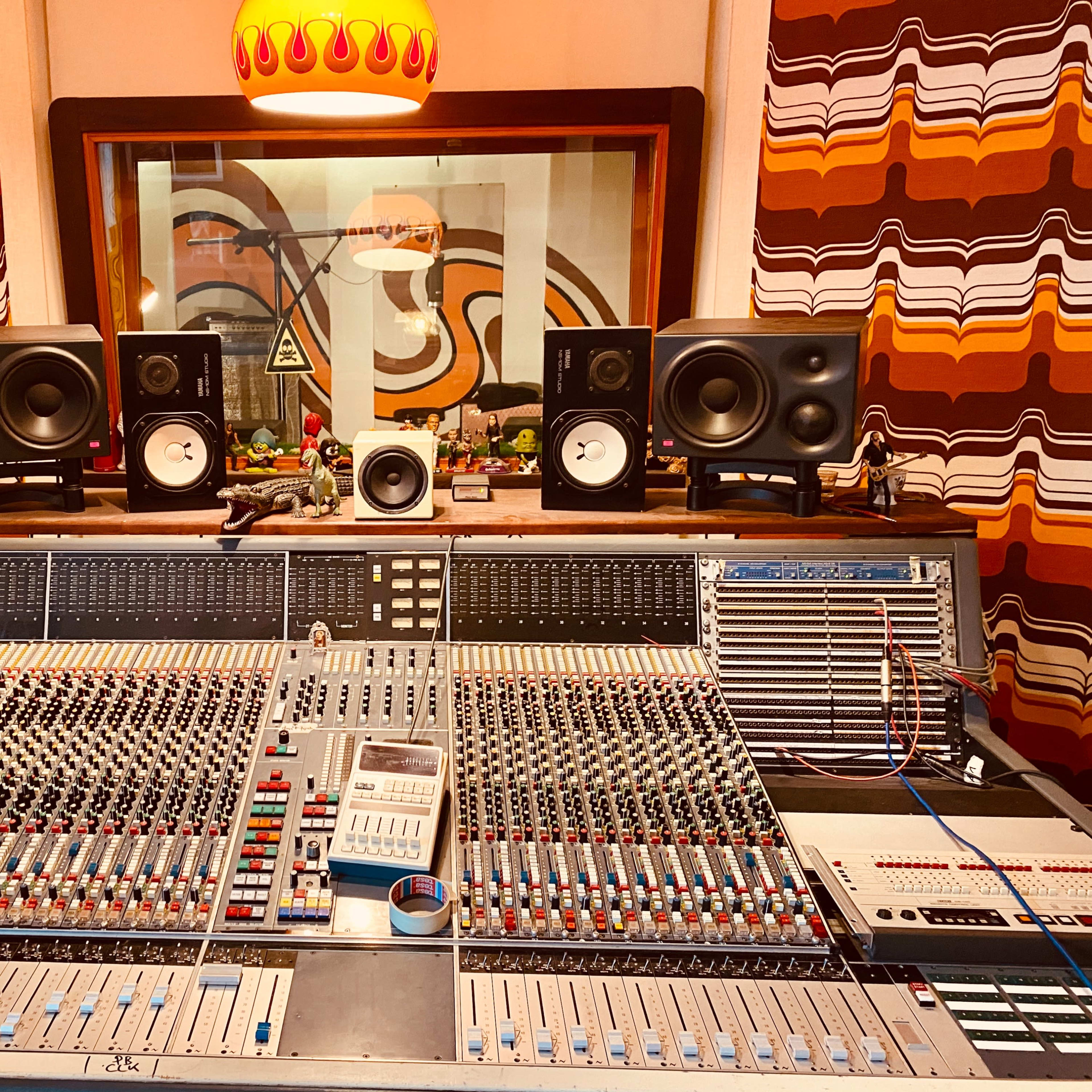 A music recording studio with a large mixing console in the foreground and speakers positioned above, framed by colorful patterned curtains in the background.