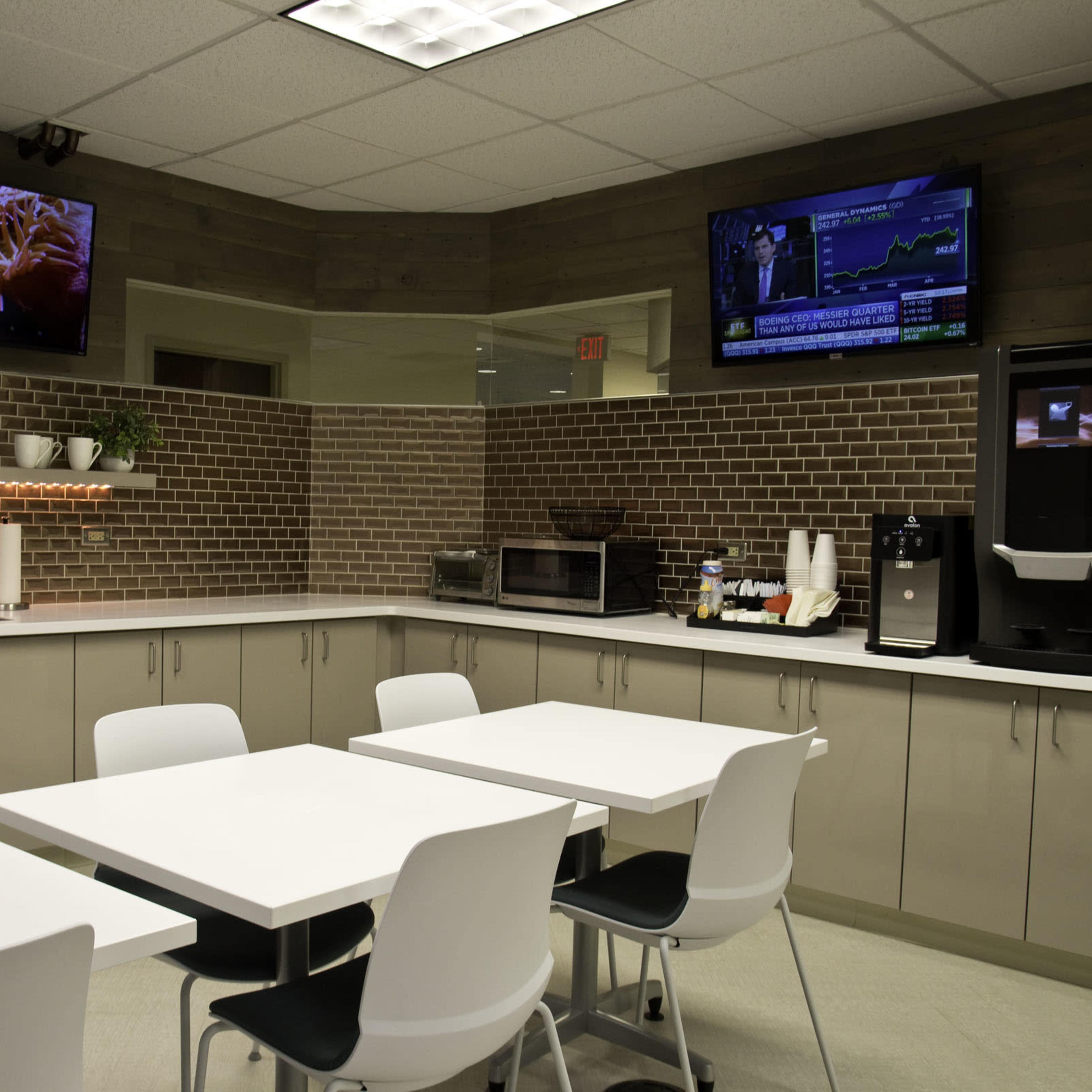 A modern break room with two televisions mounted on the walls, a kitchenette area with a refrigerator, microwave, and coffee machine, and several white tables and chairs arranged for dining.