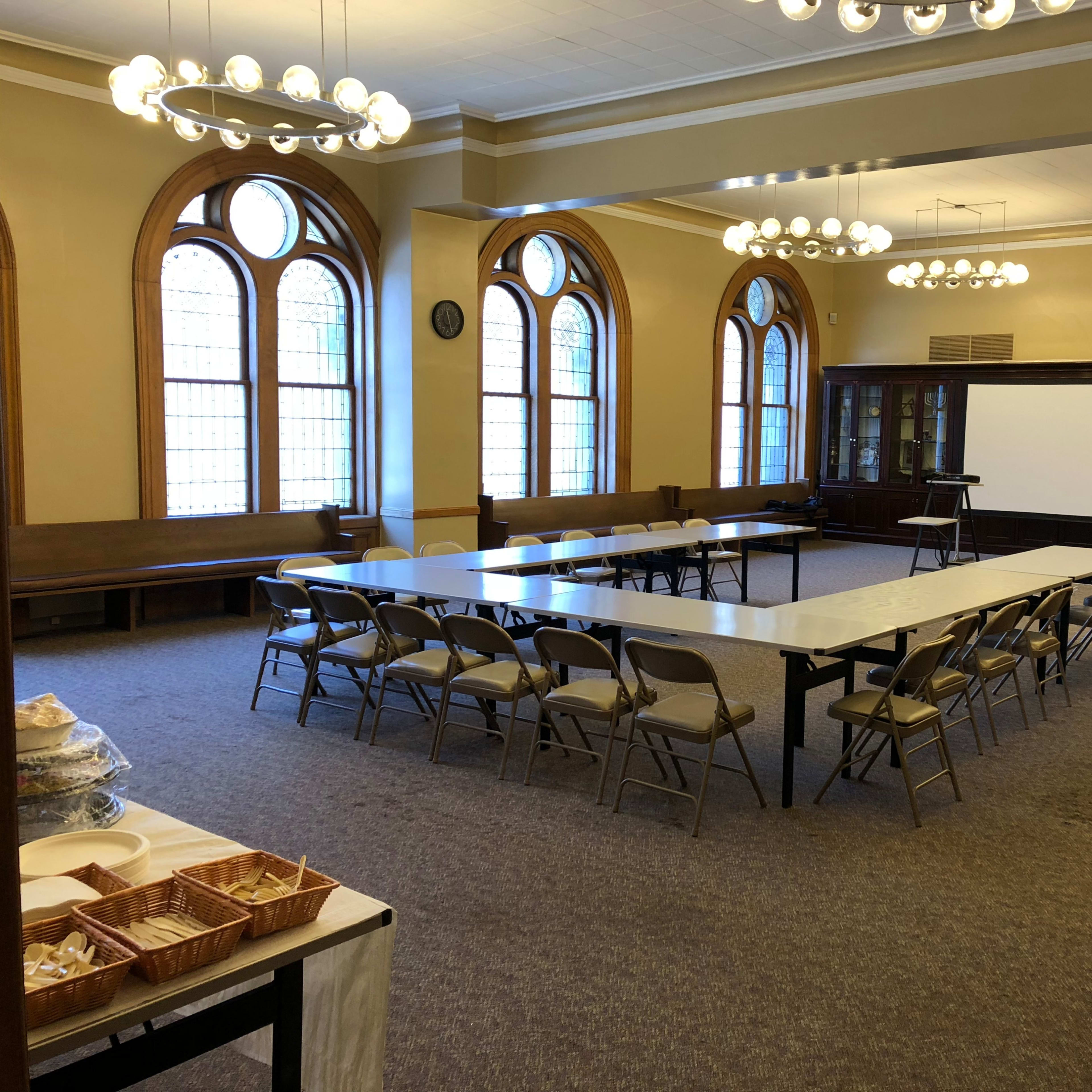 A spacious meeting room features a long table surrounded by folding chairs, with large windows and a projector screen at one end.