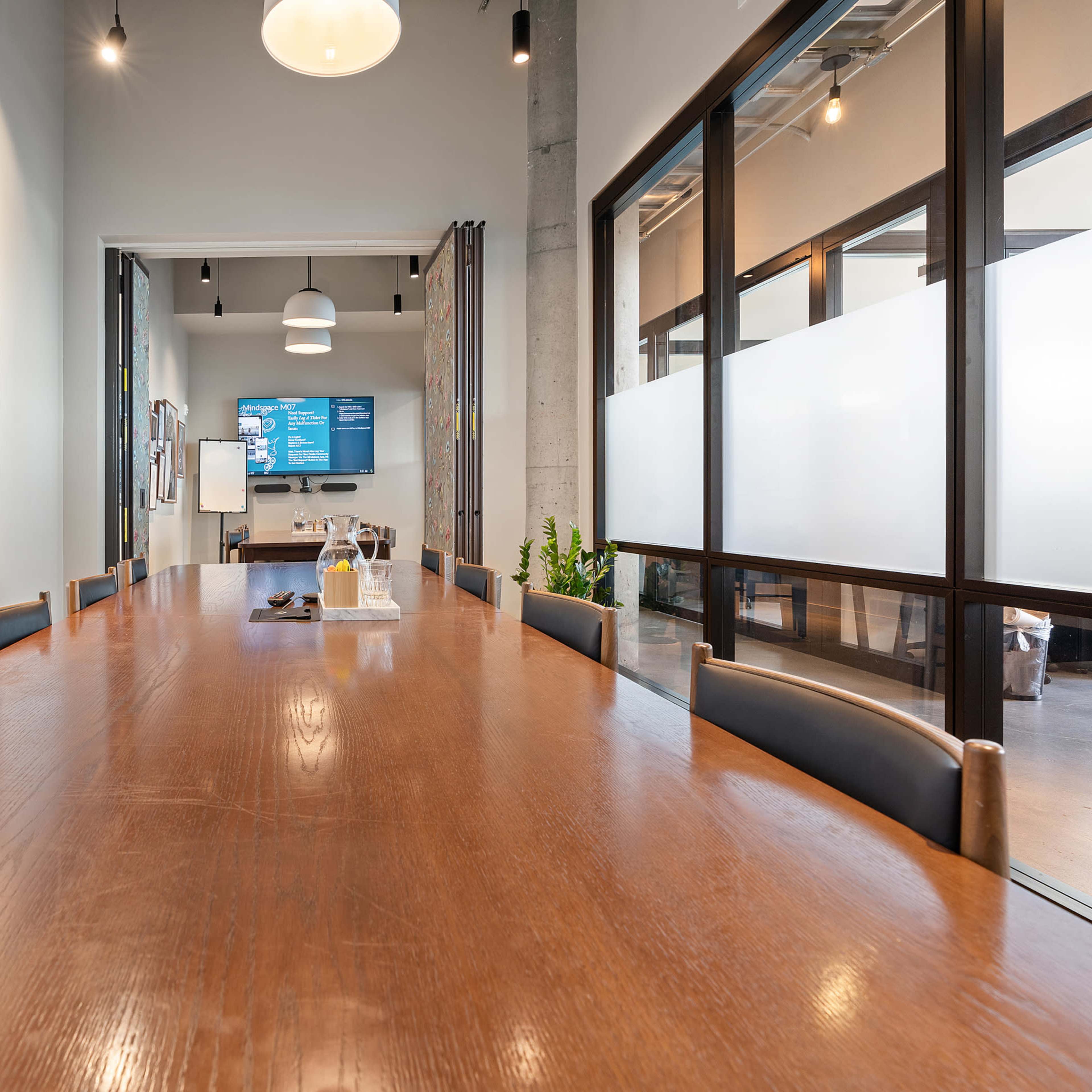 A long wooden table is set in a modern meeting room with framed art on the walls and a glass partition leading to a kitchen area.