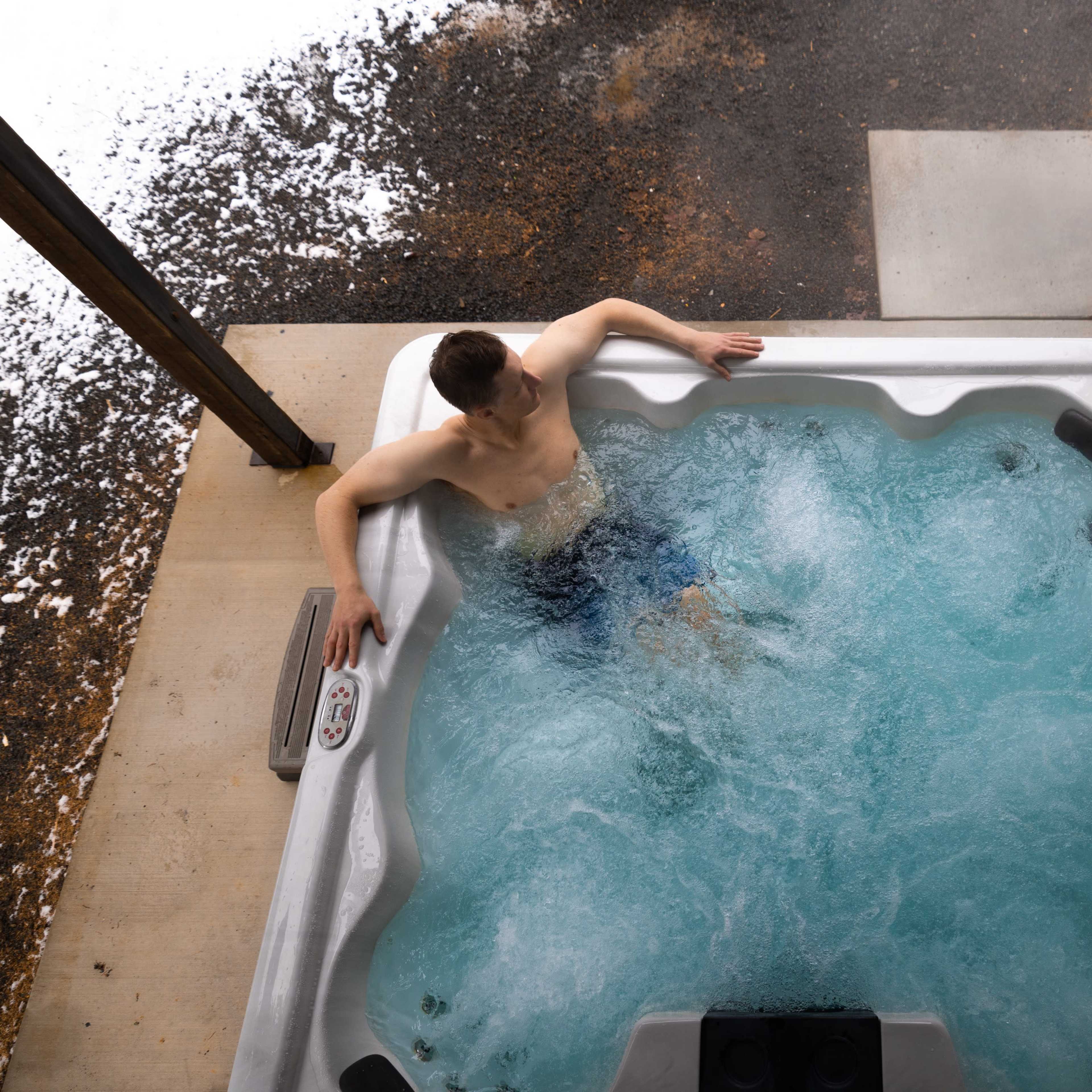 A person is sitting in a hot tub with swirling water next to a building and a snow-covered ground.