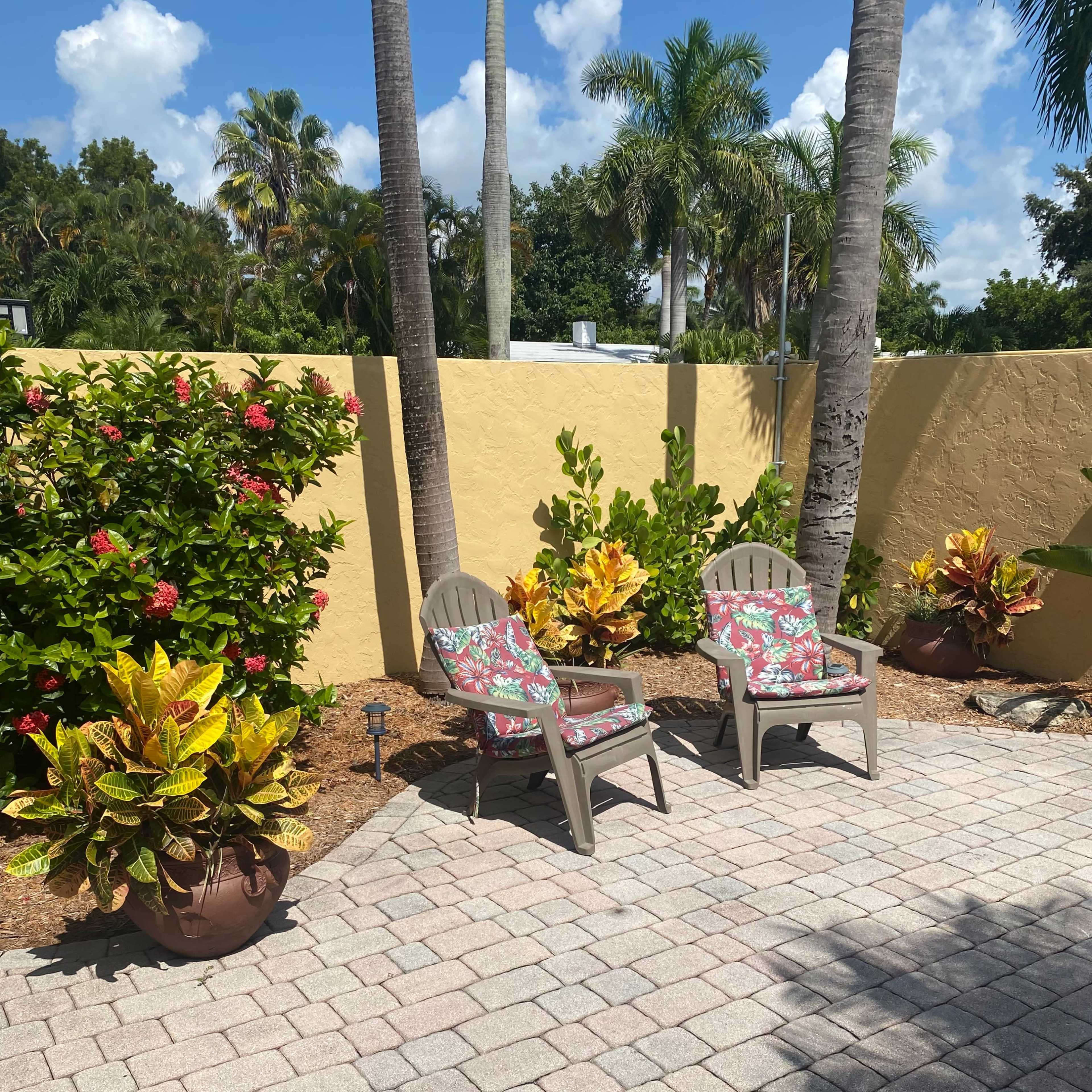 Two lawn chairs with floral cushions positioned on a paved patio surrounded by colorful plants and a yellow wall.
